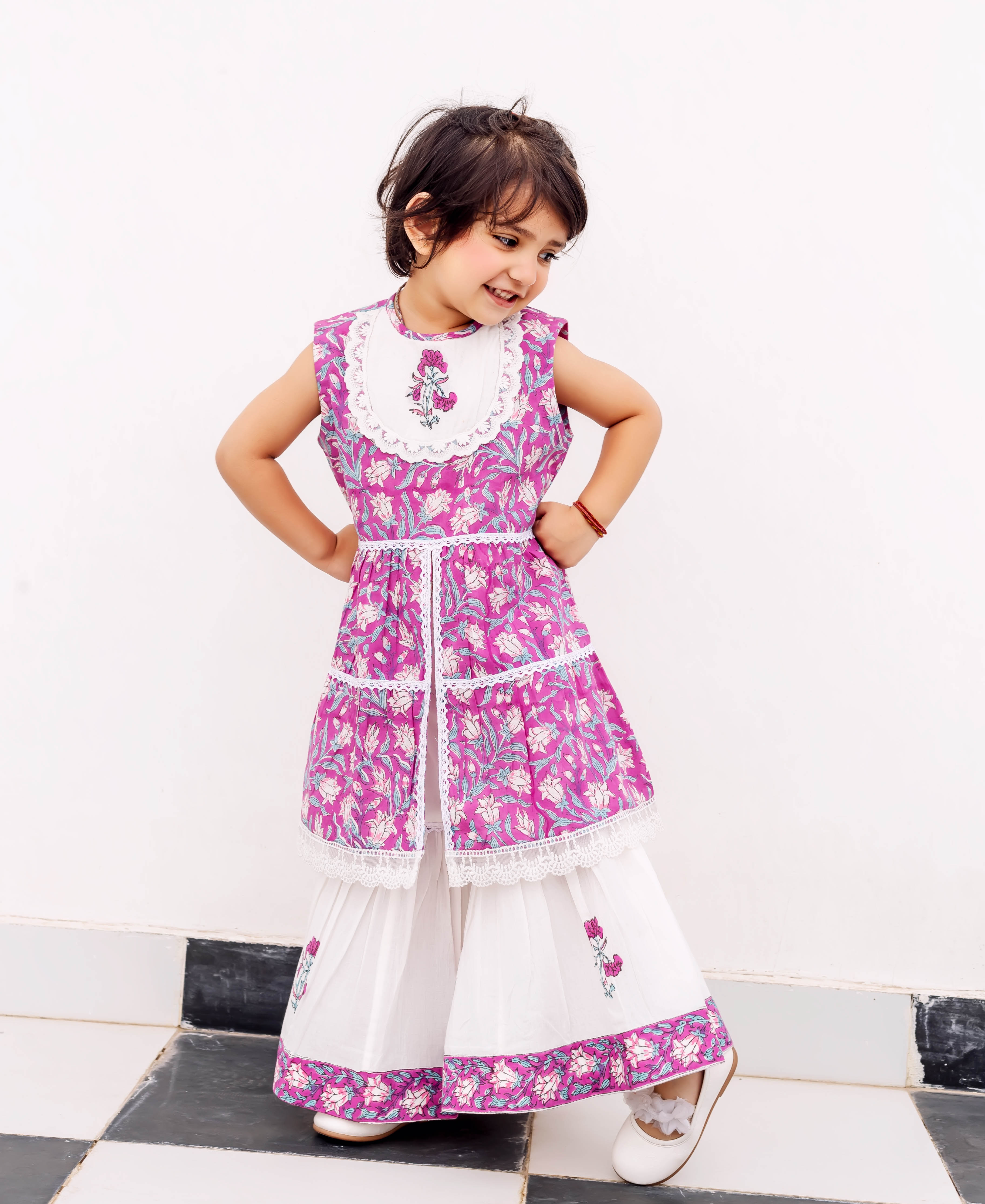 Smiling girl in purple hand-block printed kurta with white sharara and floral patterned borders.