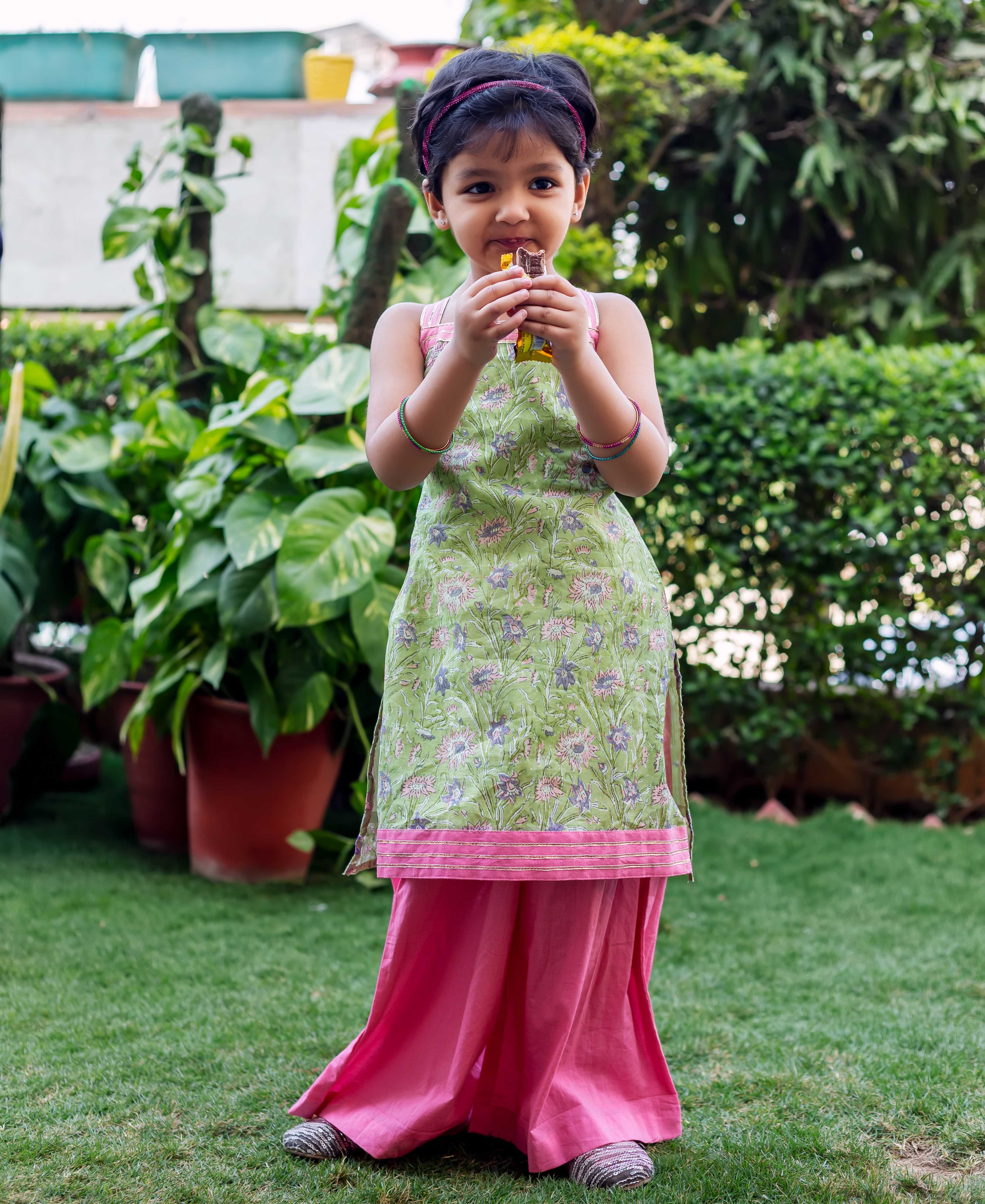 Child model wearing a green hand-block-printed kurta with pink sharara pants, standing outdoors.