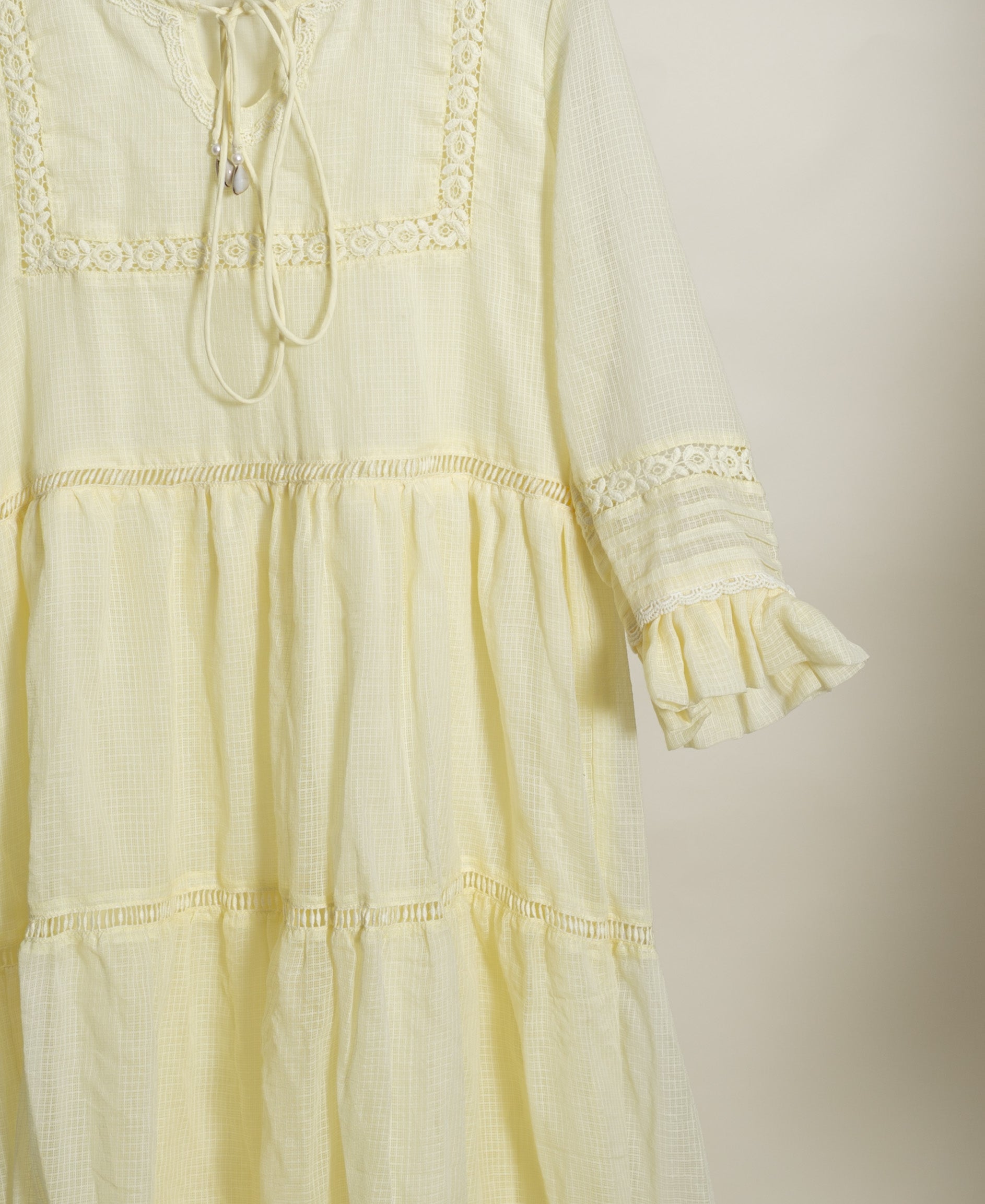 Flat lay of pale yellow tiered cotton dress with woven texture, delicate lace, and drawstring.