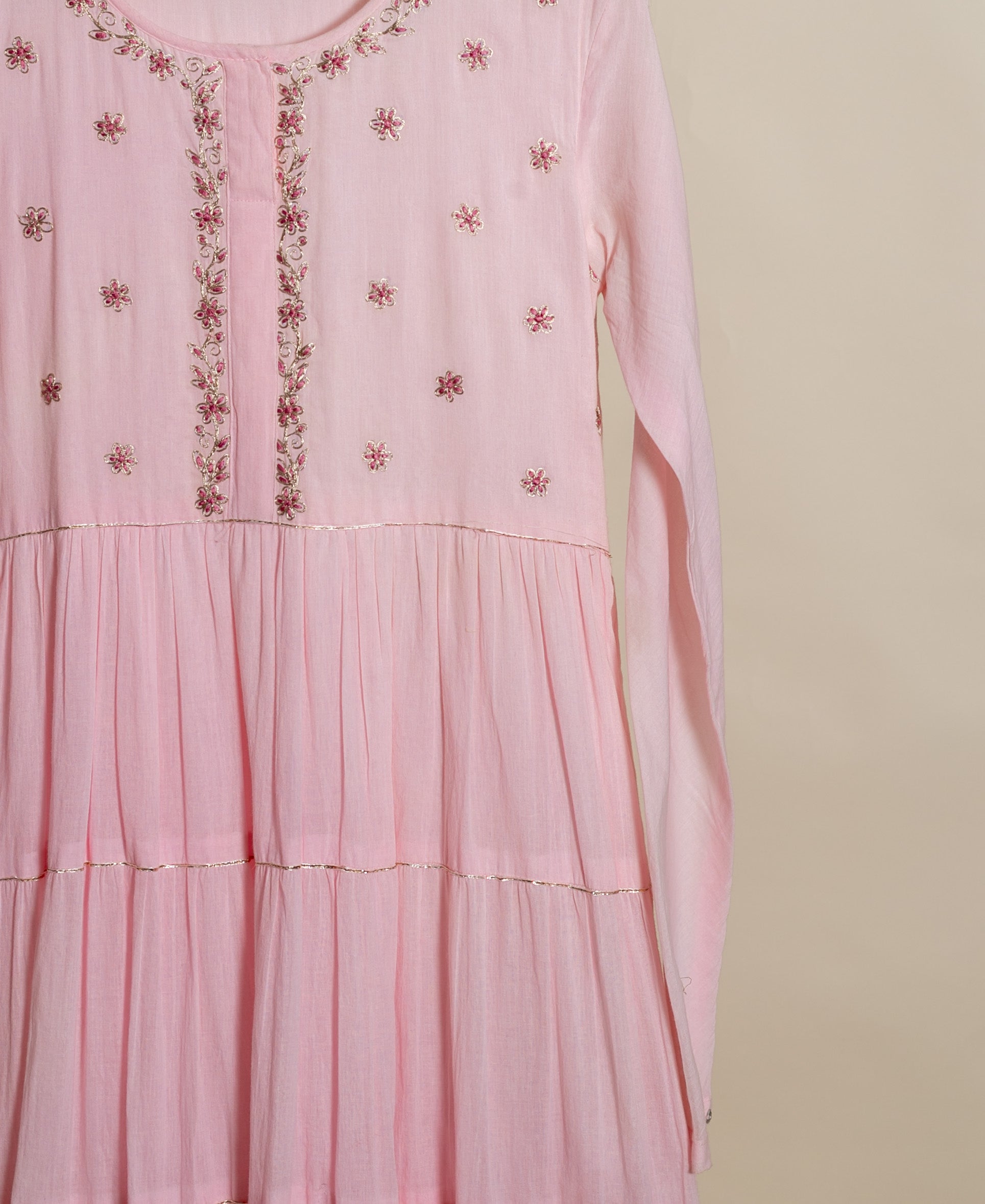 Flat lay pink tiered dress with delicate floral embroidery, gold trim, and long sleeves.