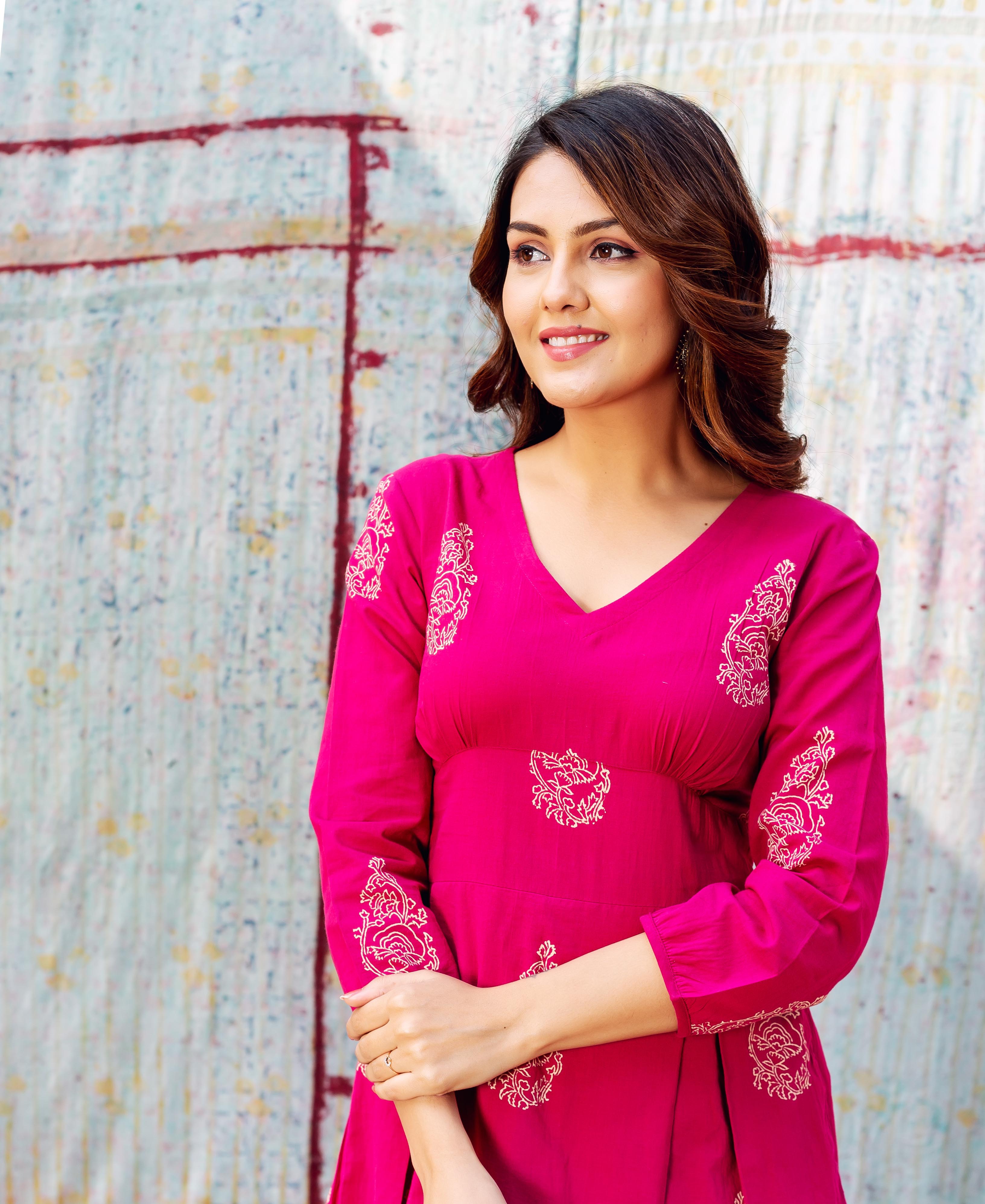 A woman in a pink dress with white floral print, standing in front of a wall.