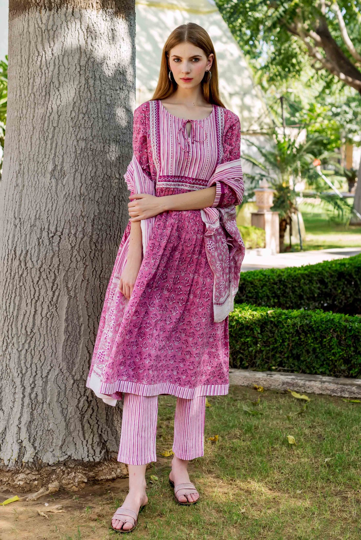 Model standing, wearing a pink hand-block printed A-line kurta, striped pants and matching dupatta.