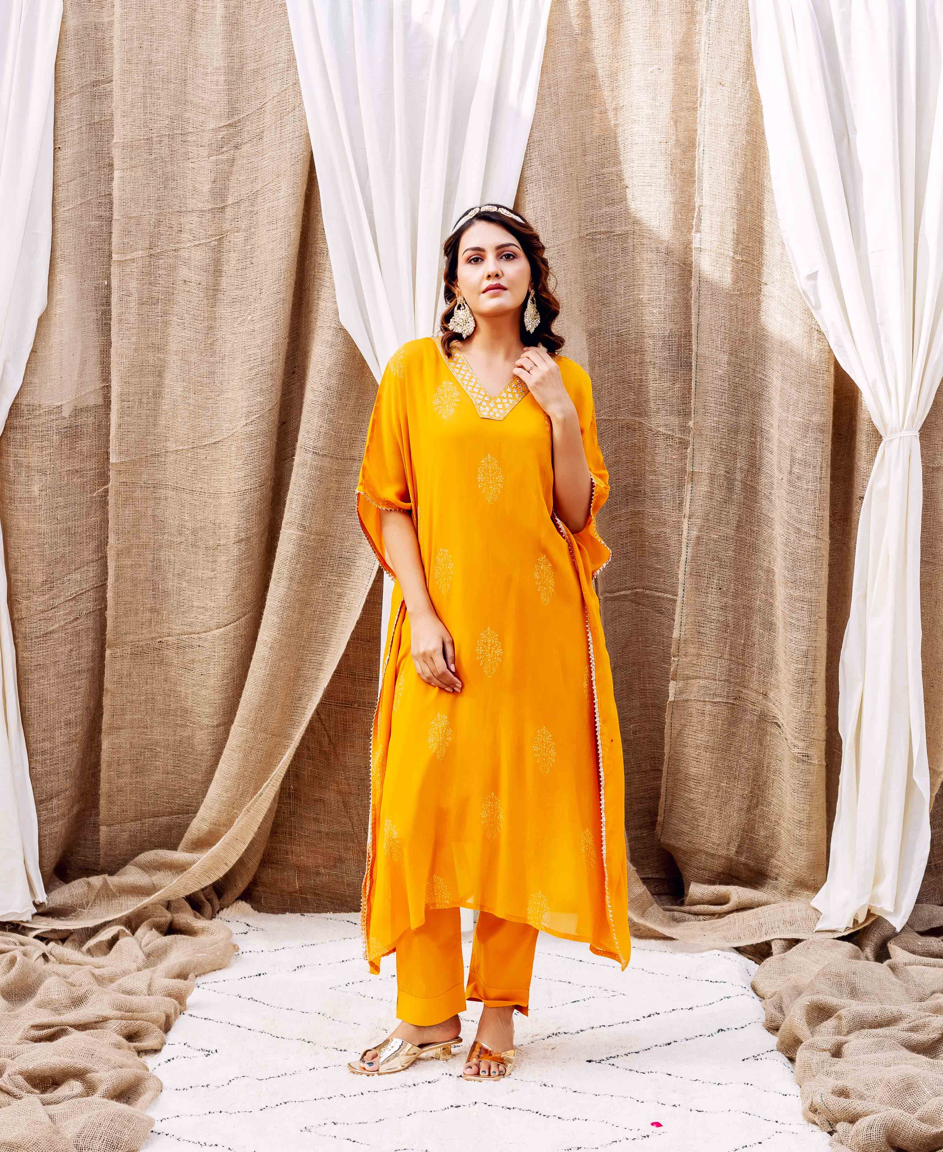 Model wears yellow embroidered kaftan set, standing against white and burlap drapes.