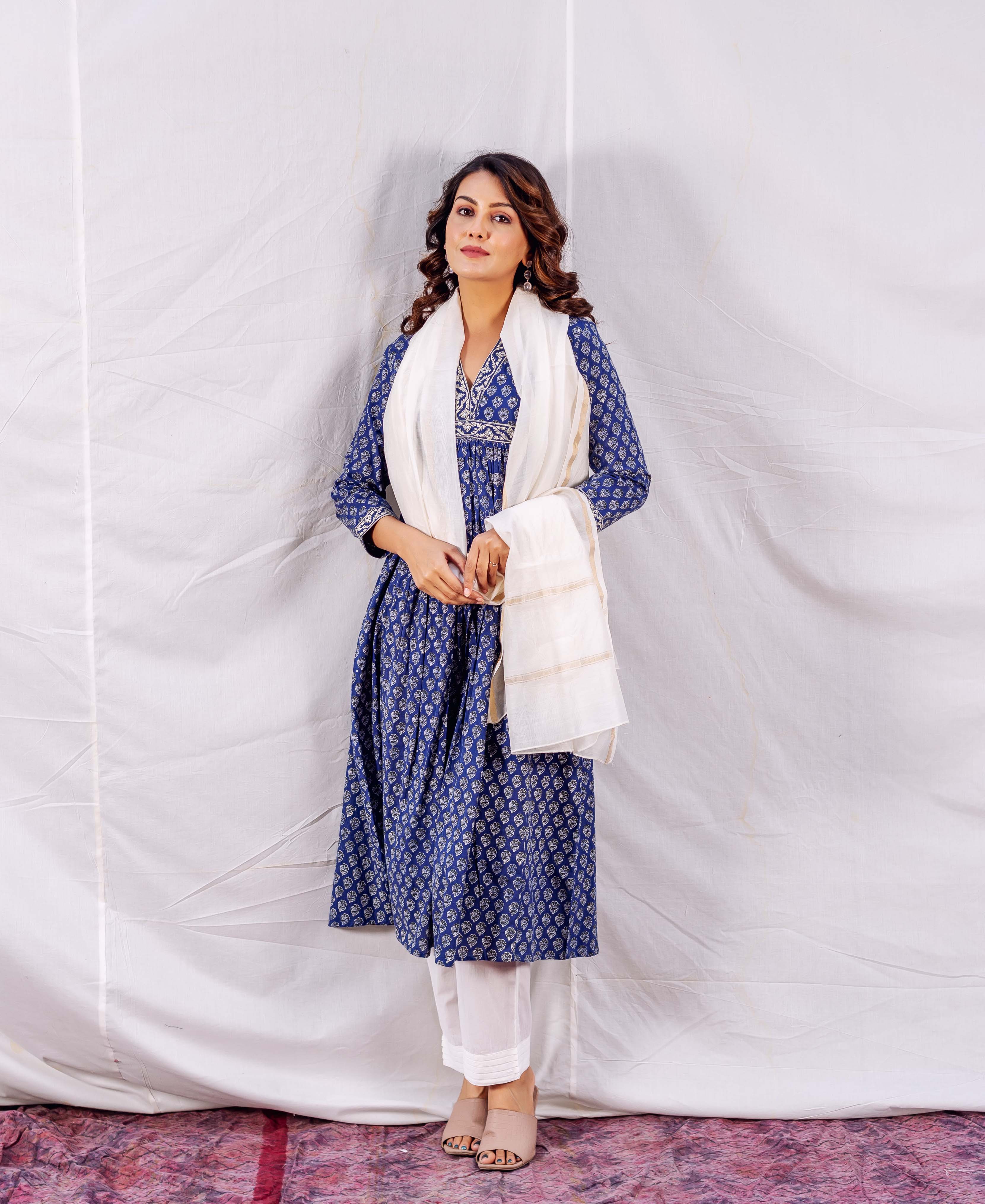 Woman wearing blue patterned dress and white scarf and pants, standing against a white sheet background.