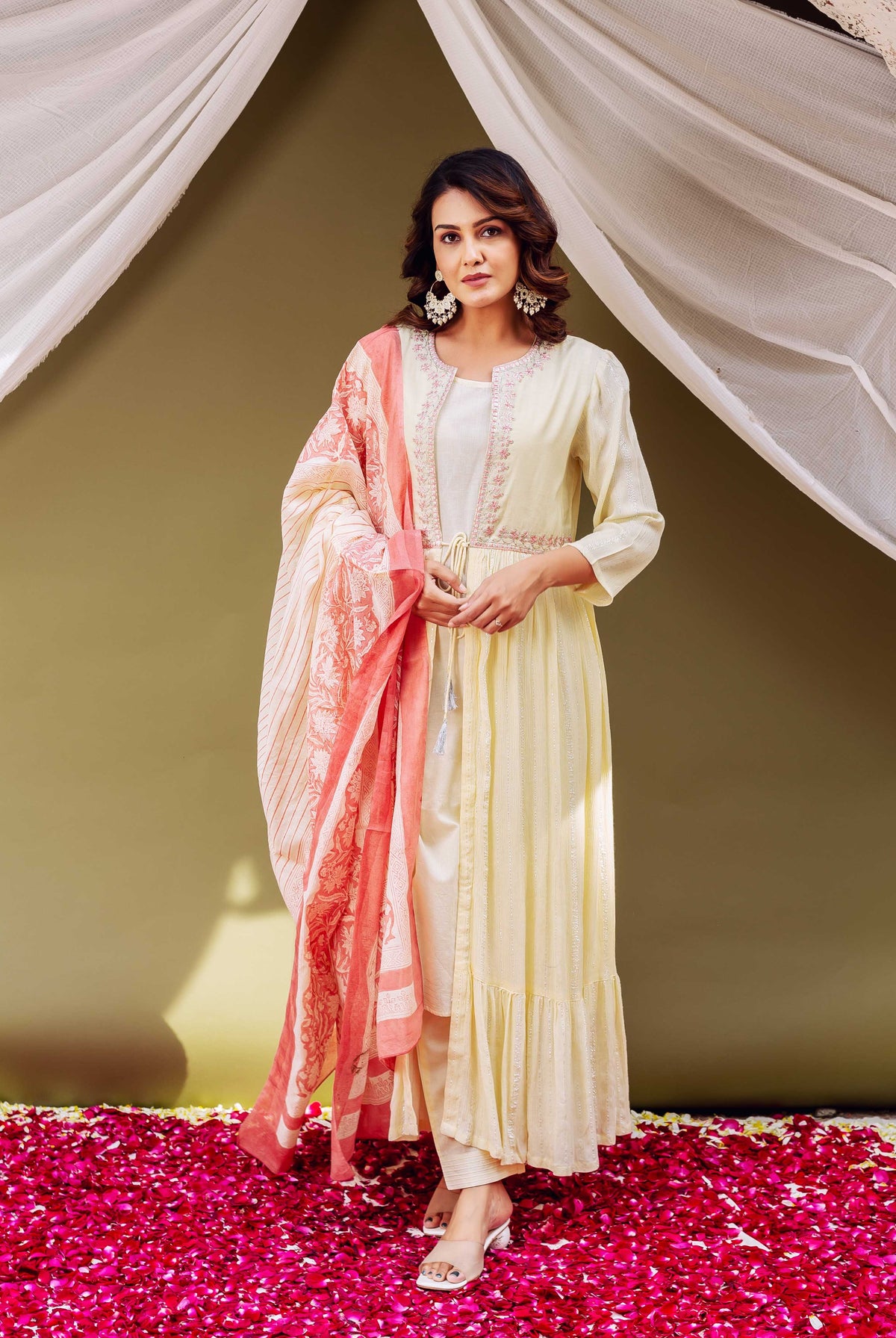 Woman in cream kurta with embroidered waist and pink and white shawl standing on flower petals.