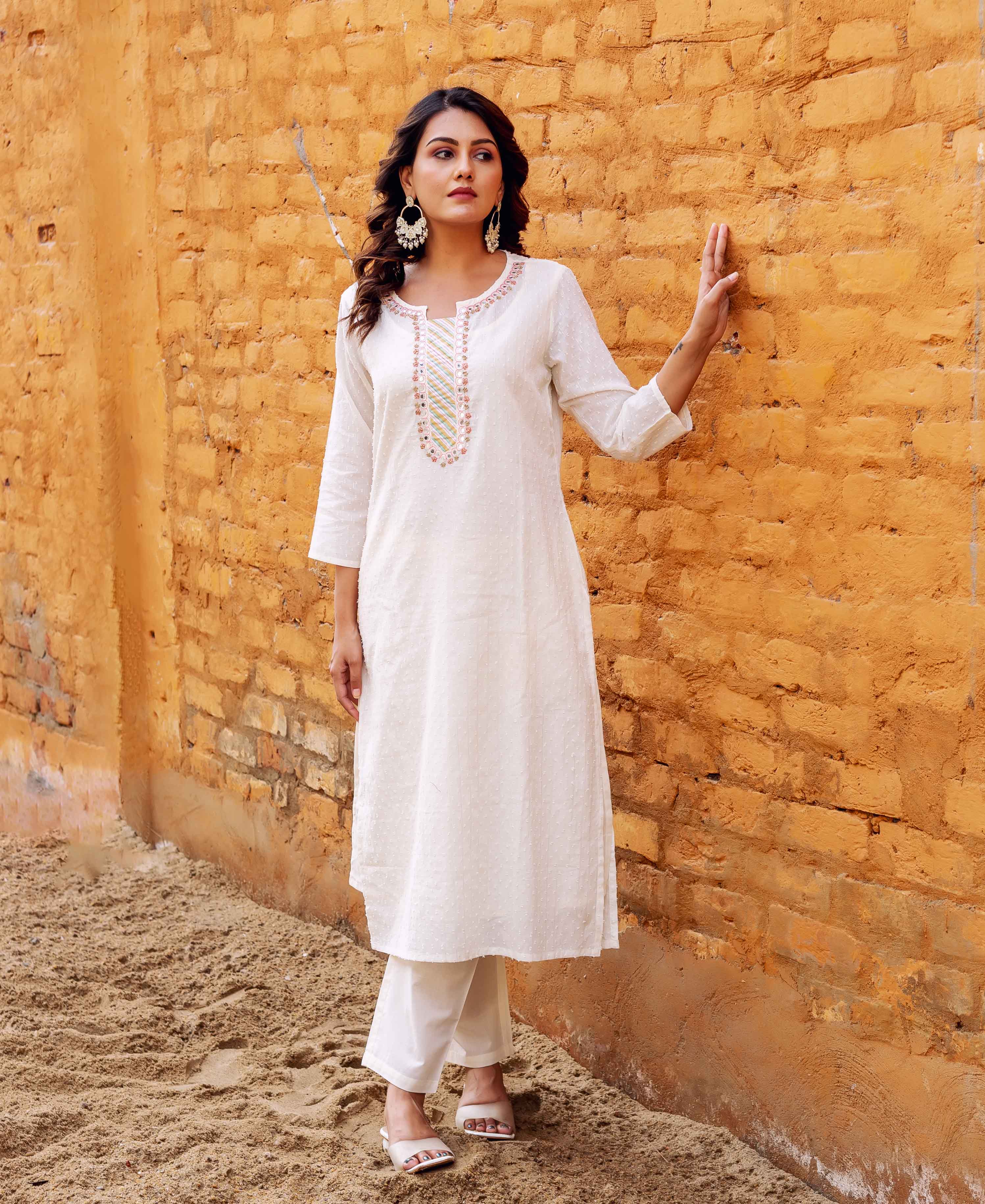 A woman in a white kurta with intricate block-printed designs stands against a brick wall.