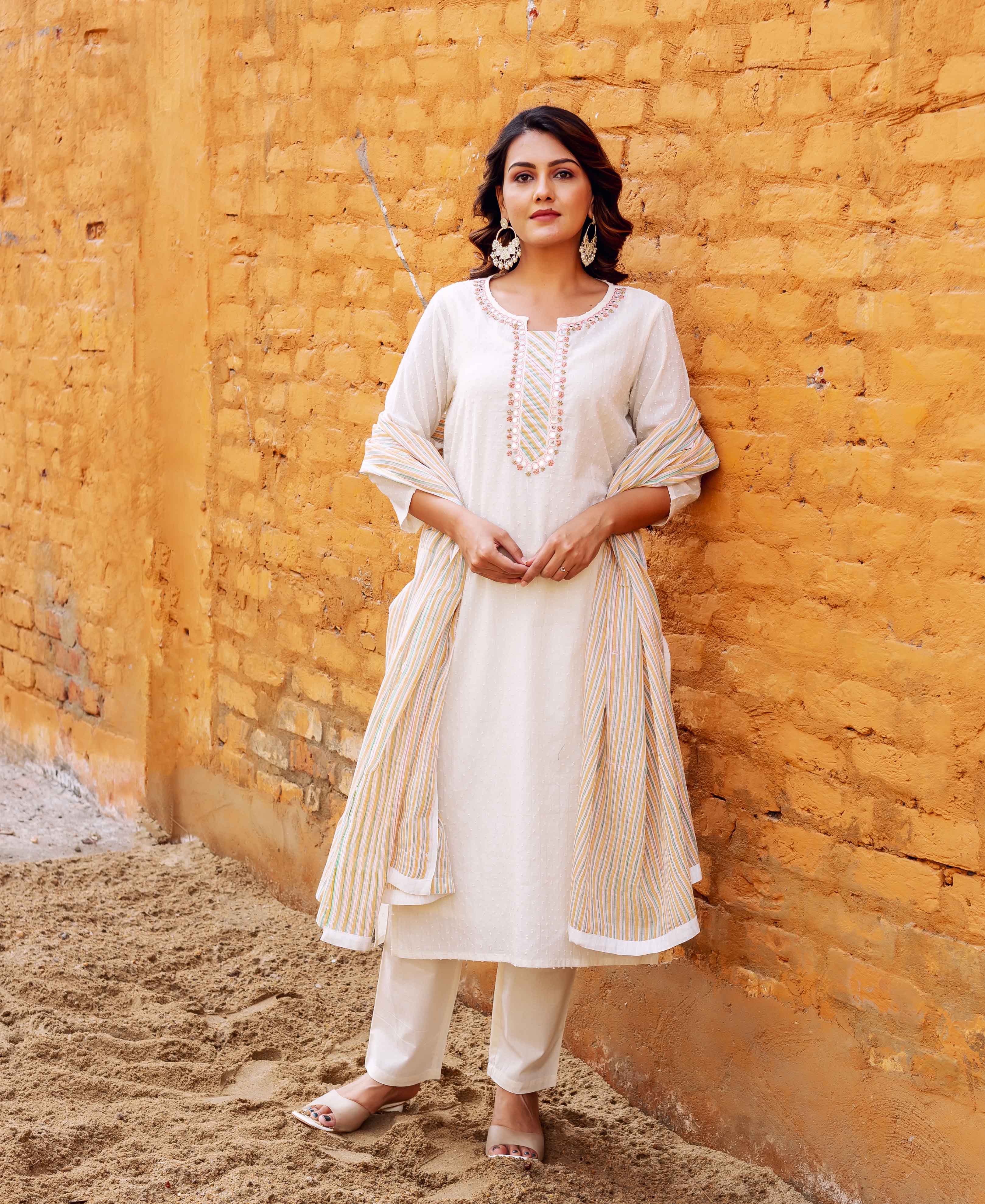 A woman in a white kurta with a striped dupatta, standing against a brick wall.