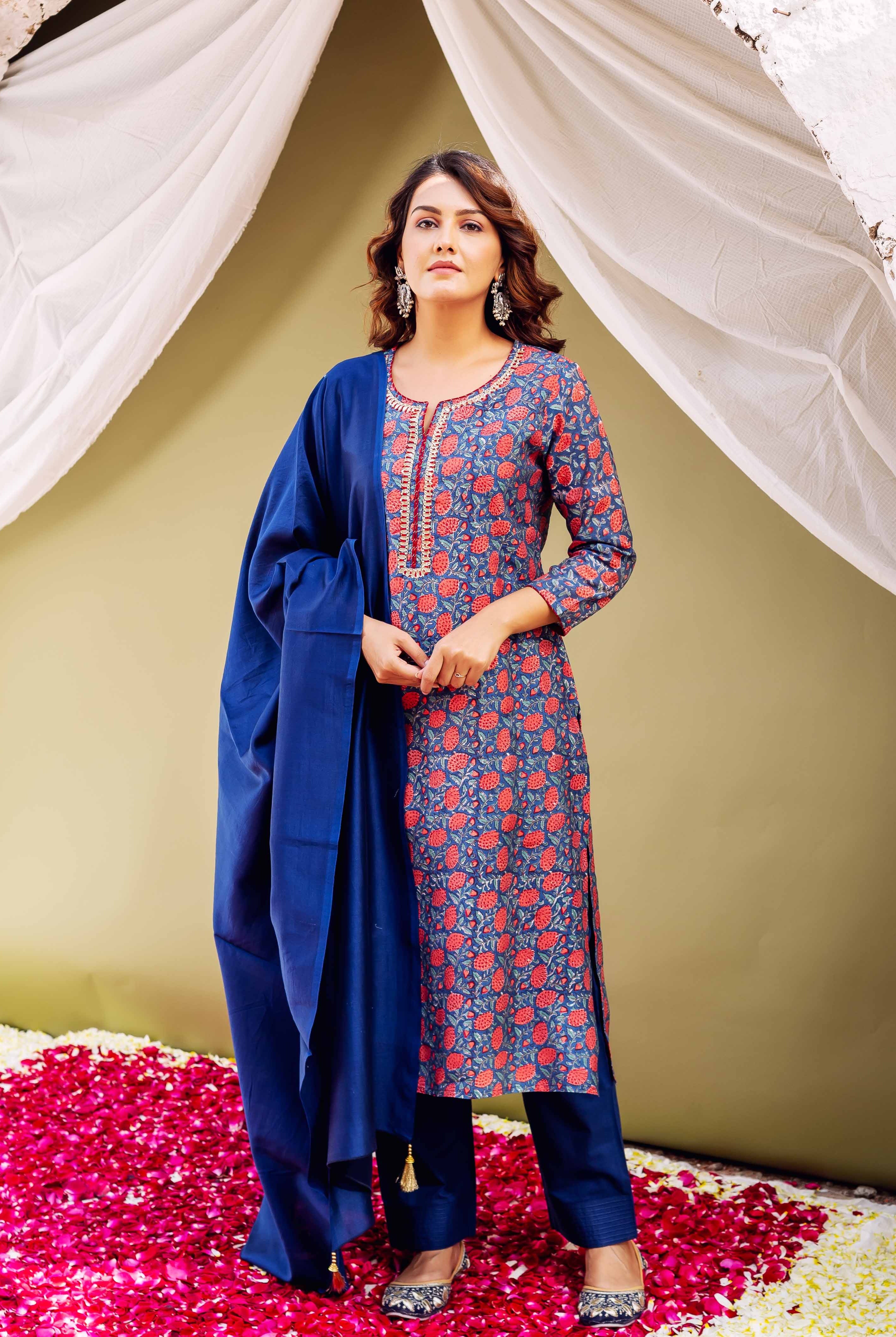 A woman in a blue and red floral kurta with gold accents, standing on rose petals.