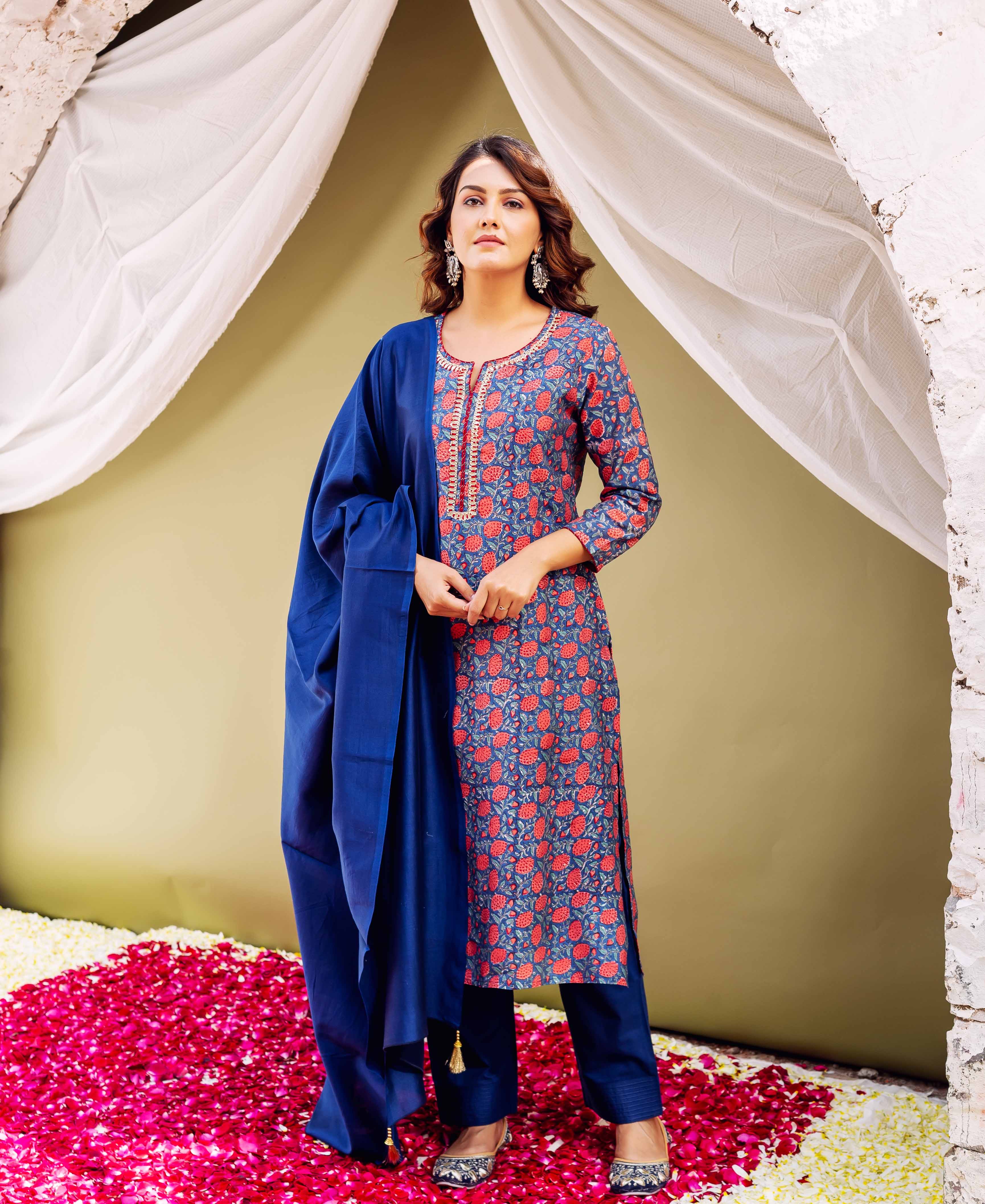 A woman in a blue and red floral kurta with gold accents, standing on rose petals.