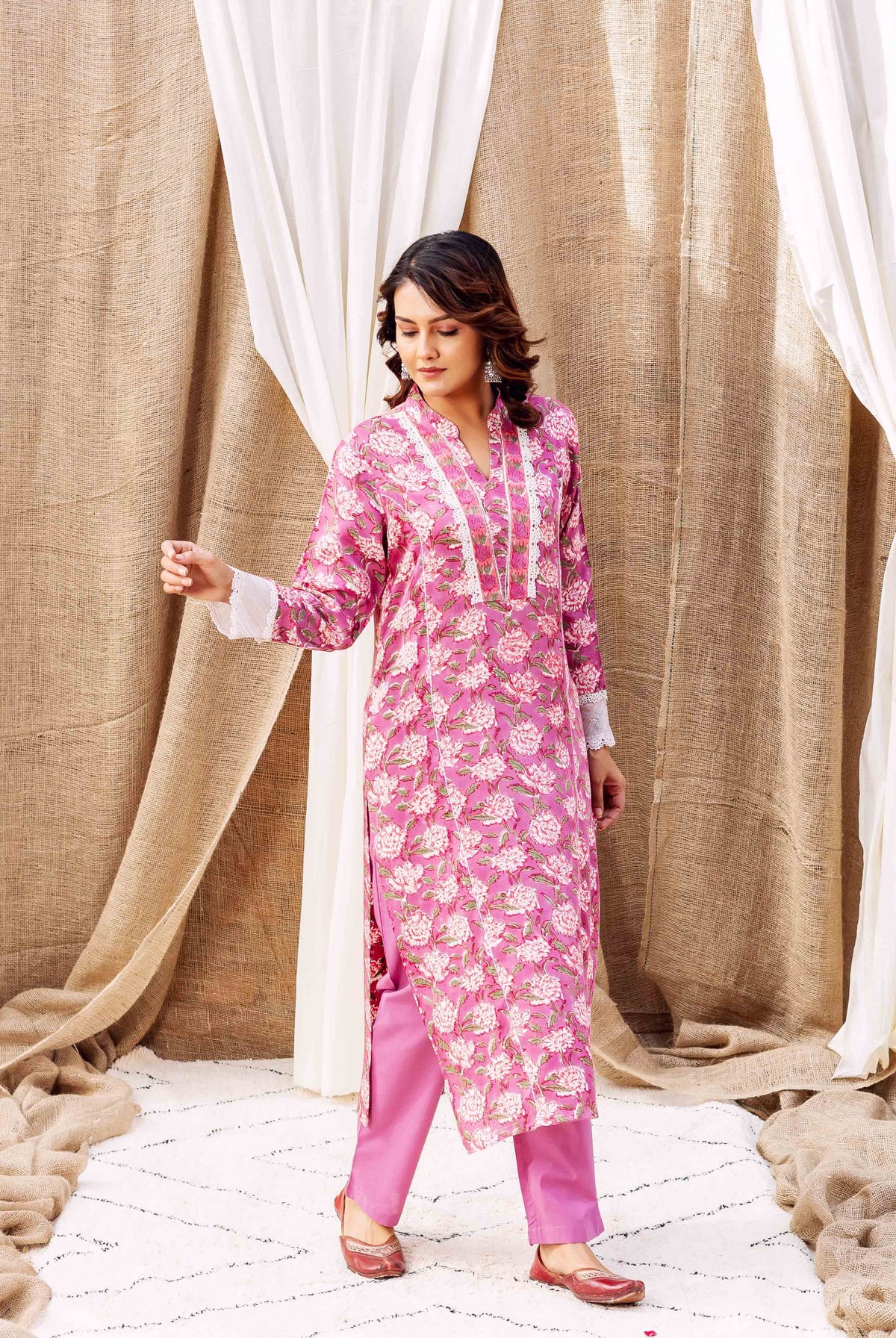 Woman models pink floral hand-block printed kurta with white lace trim and matching pants.