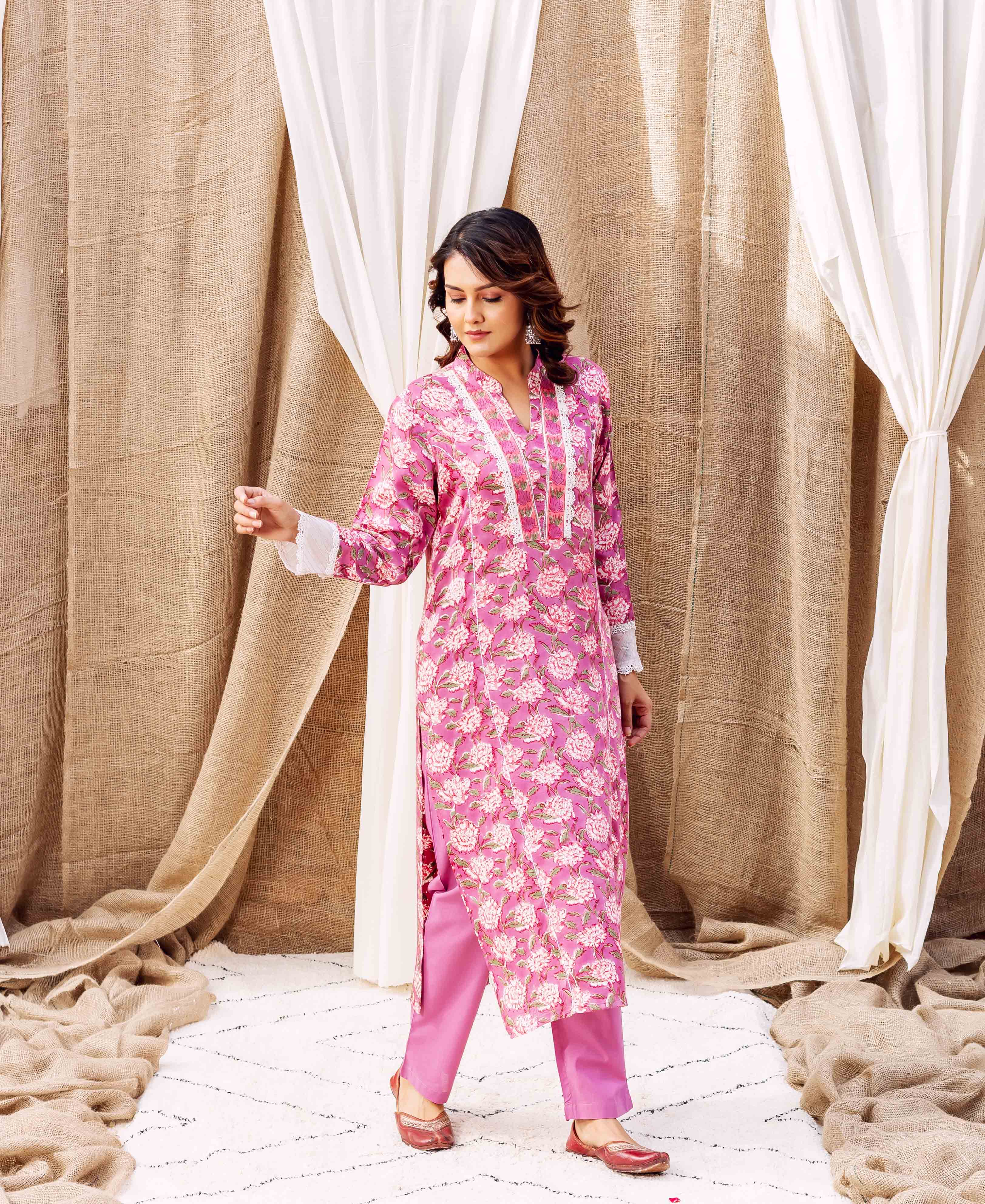 Woman models pink floral hand-block printed kurta with white lace trim and matching pants.