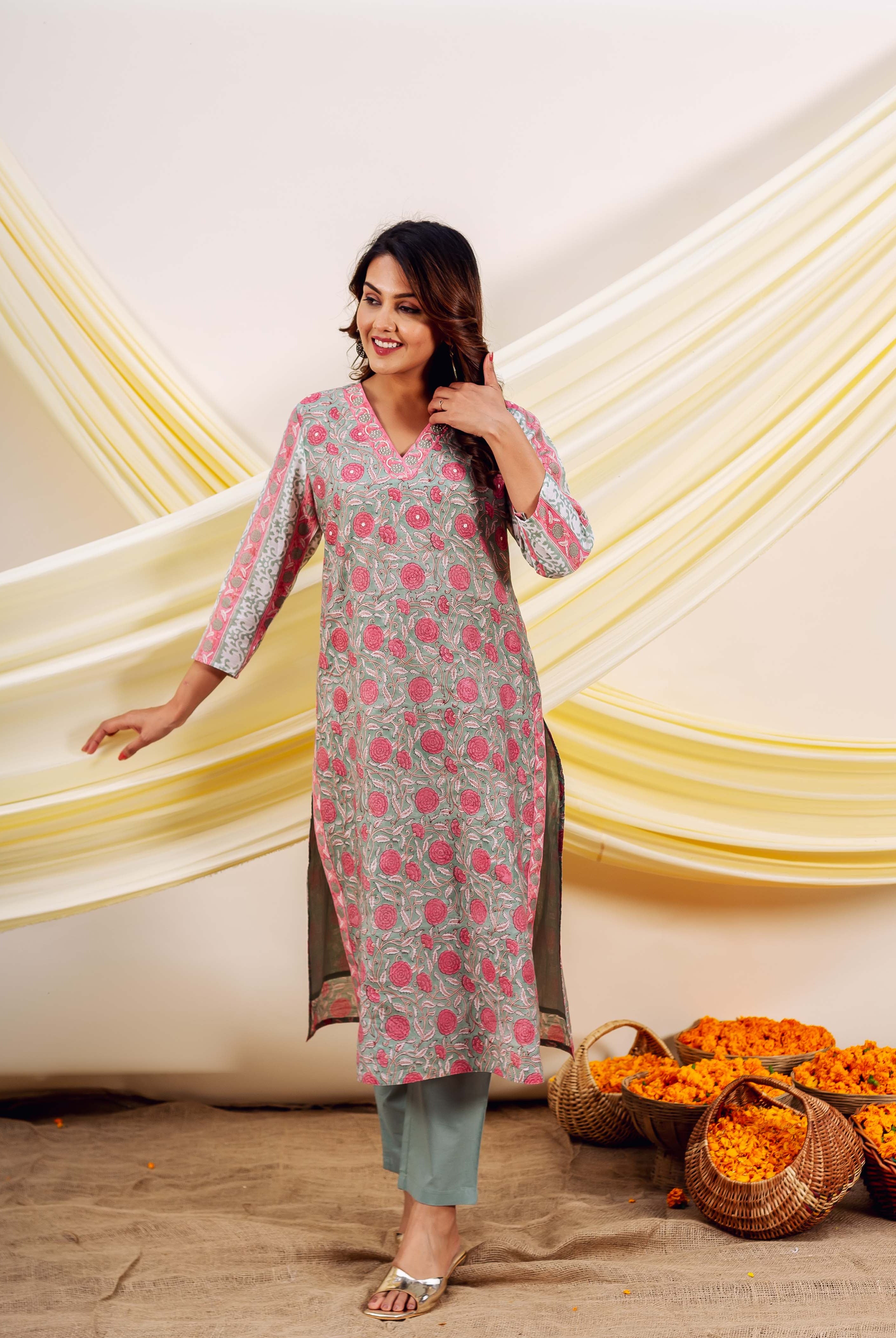 Smiling woman in a grey block-printed kurta with pink flowers, green pants, and gold sandals.