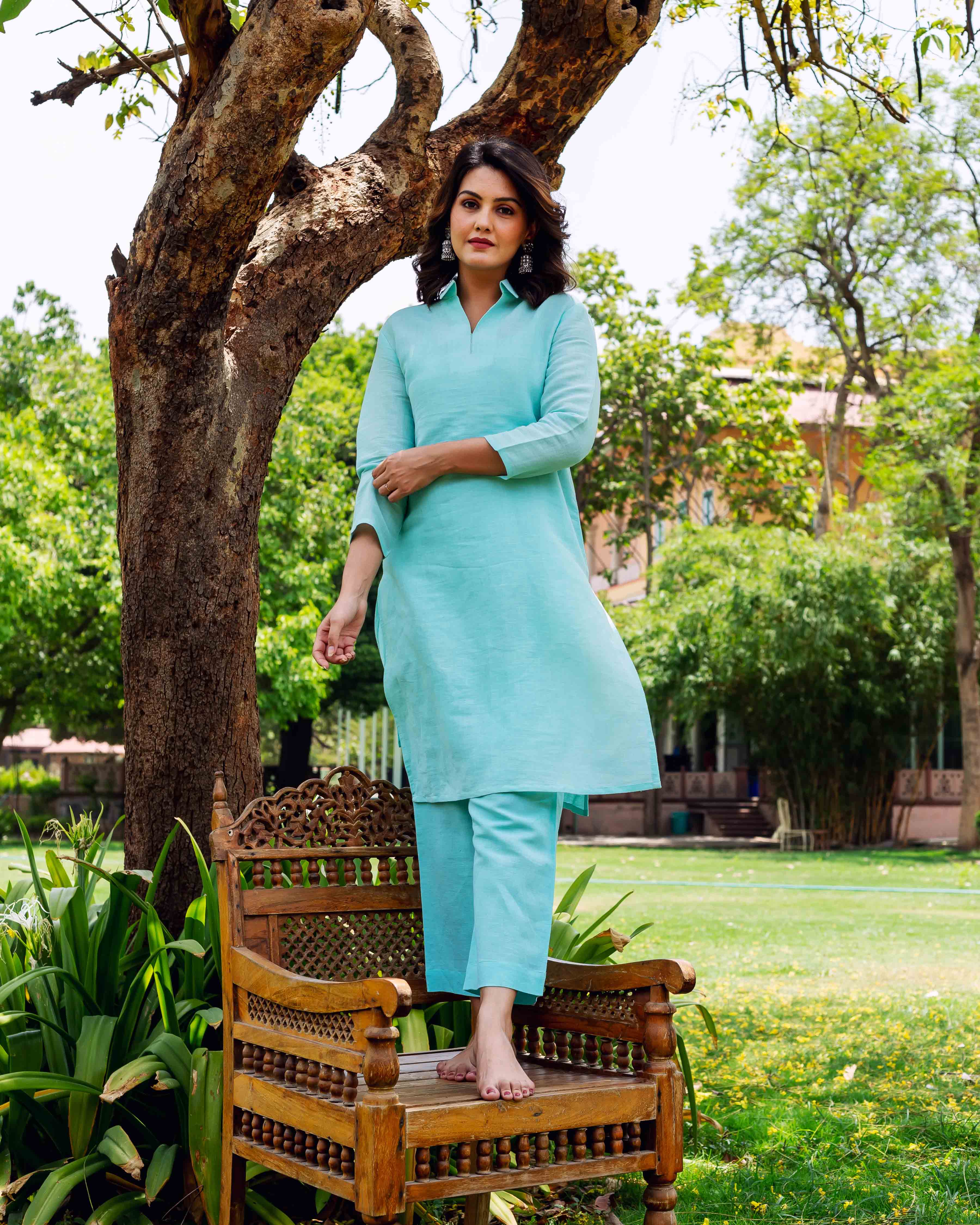 A woman standing on a wooden chair, wearing a light blue linen co-ord set.
