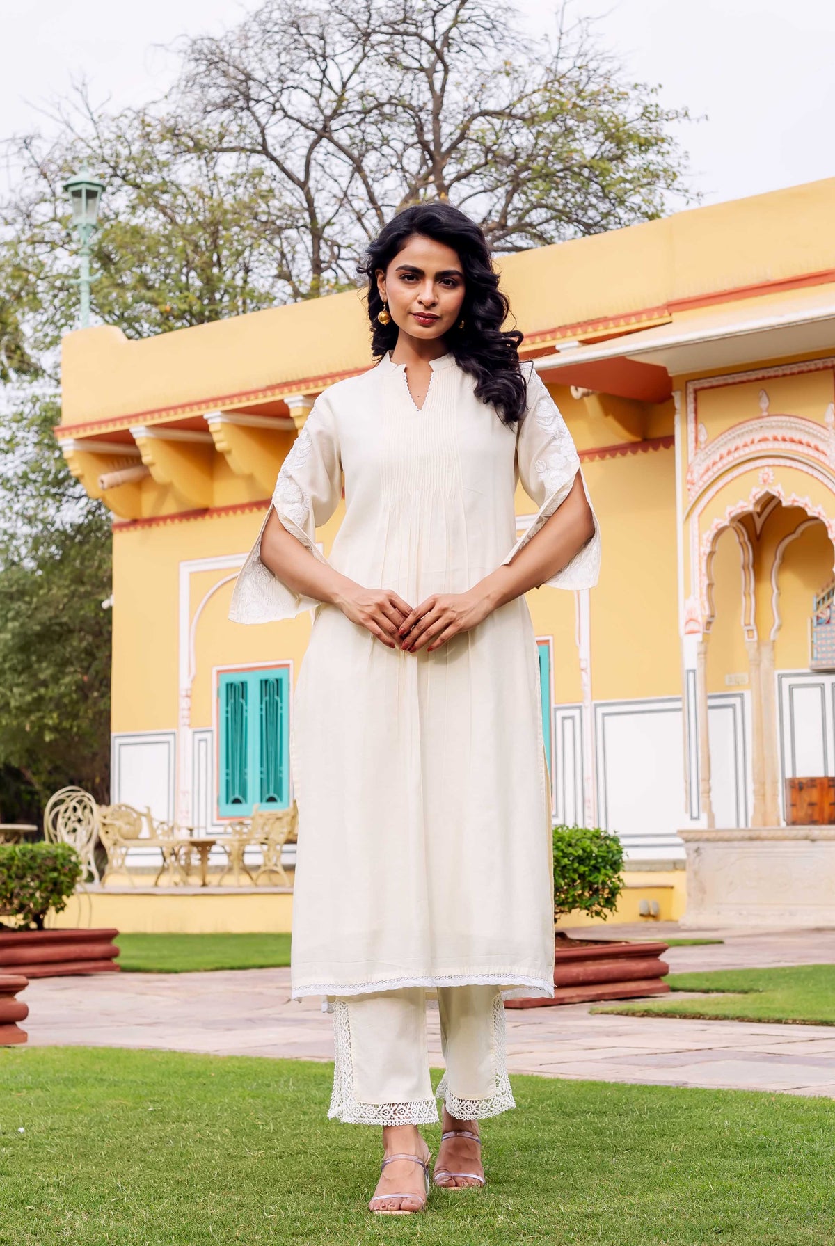 Model wearing antique white pleated kurta set with embroidered split sleeves and lace hems.