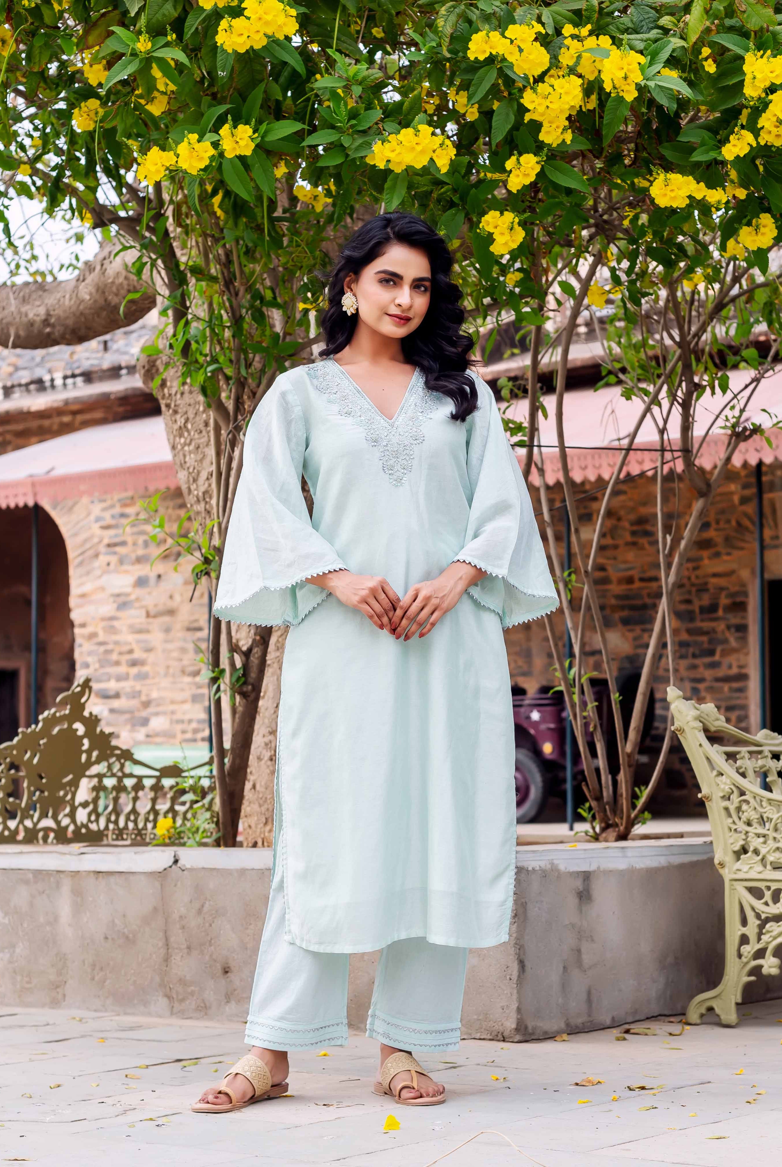 Woman in long, light blue kurta with embroidered V-neck and bell sleeves, standing outdoors.