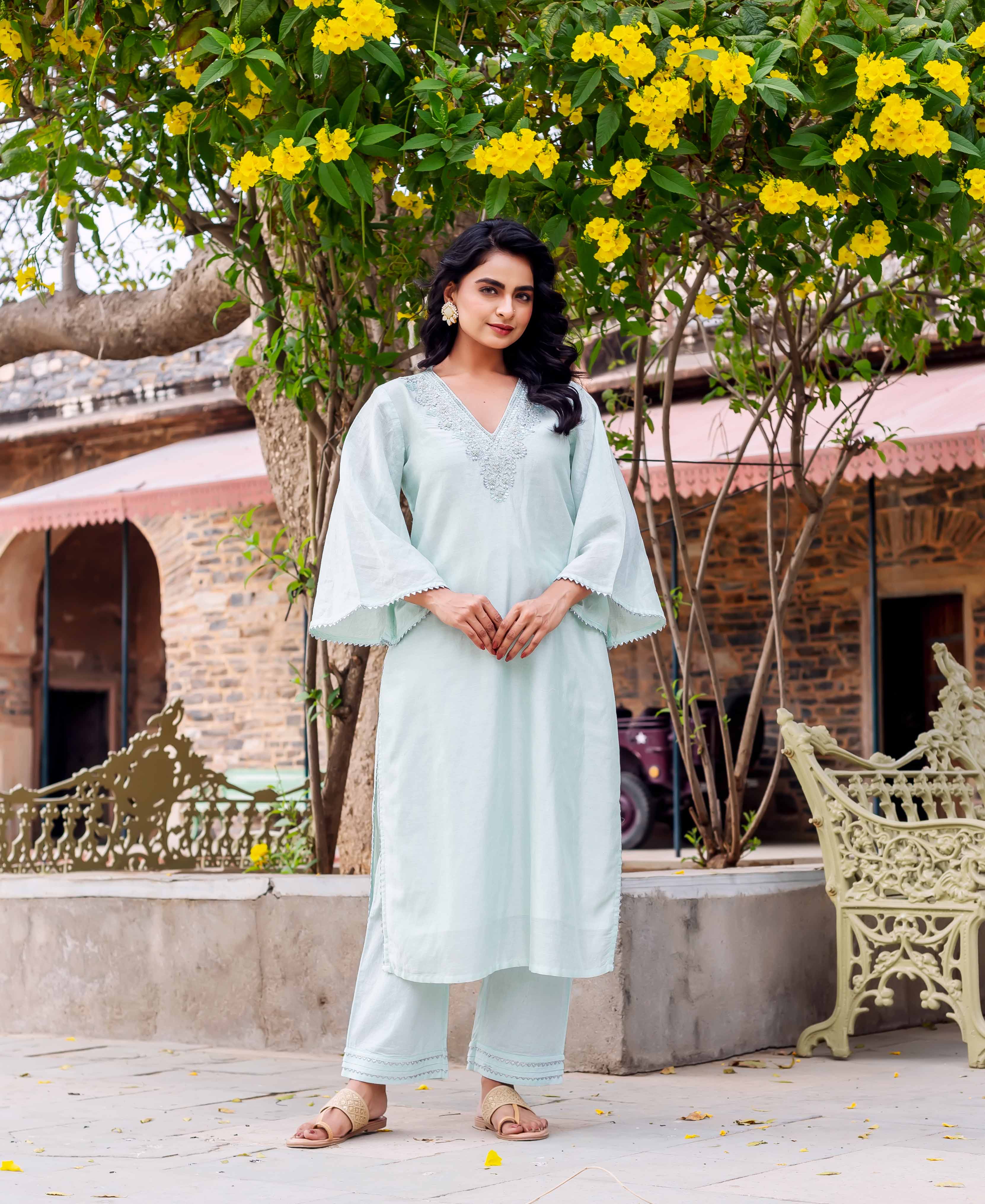 Woman in long, light blue kurta with embroidered V-neck and bell sleeves, standing outdoors.