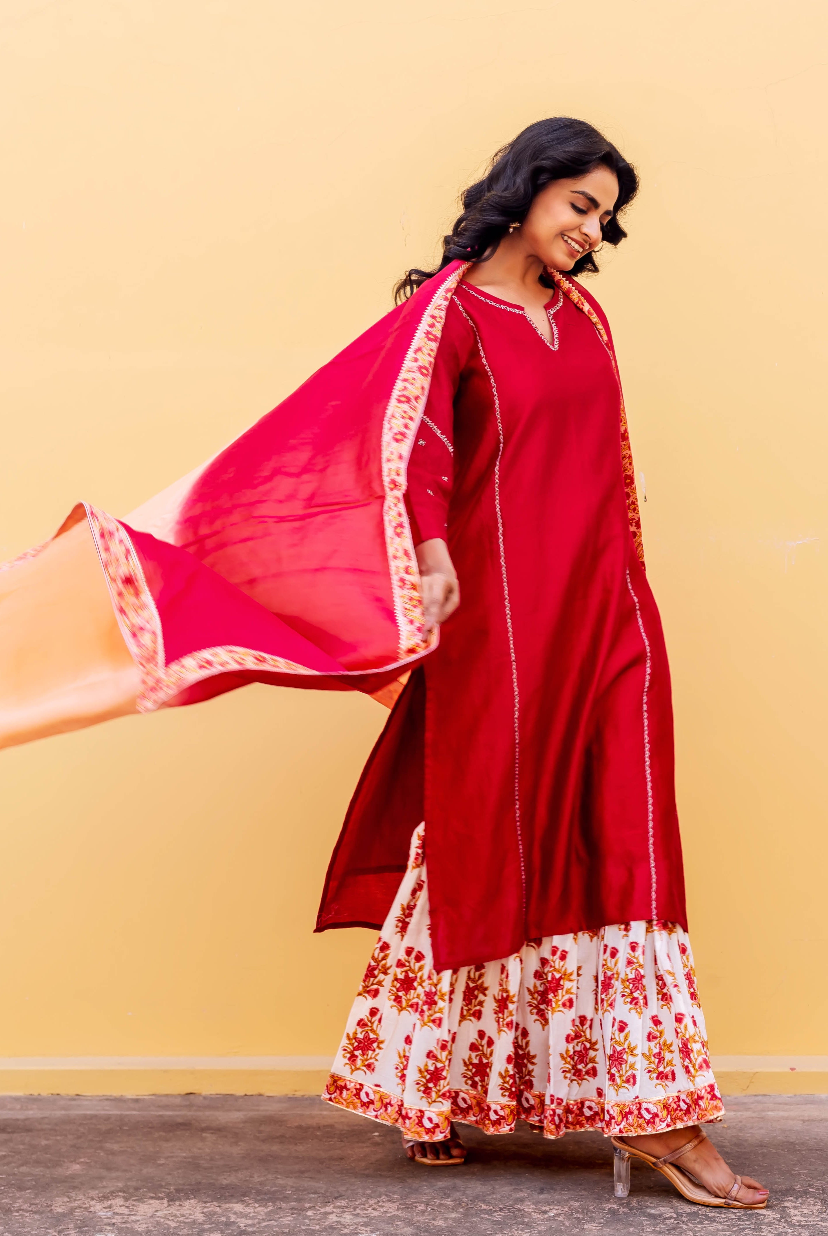 Smiling woman in red kurta, white floral hand-block print sharara, and flowing pink-orange dupatta.