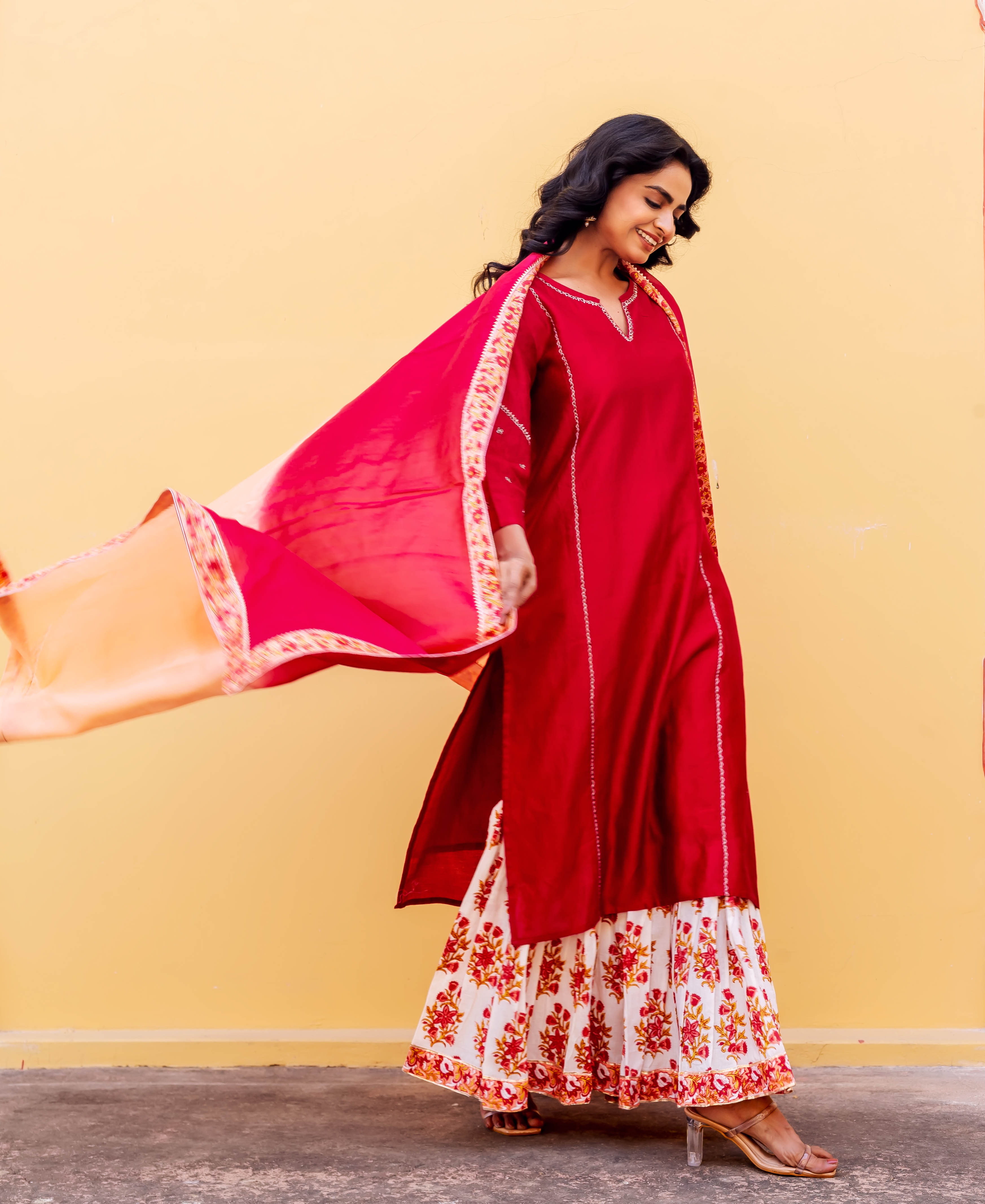 Smiling woman in red kurta, white floral hand-block print sharara, and flowing pink-orange dupatta.