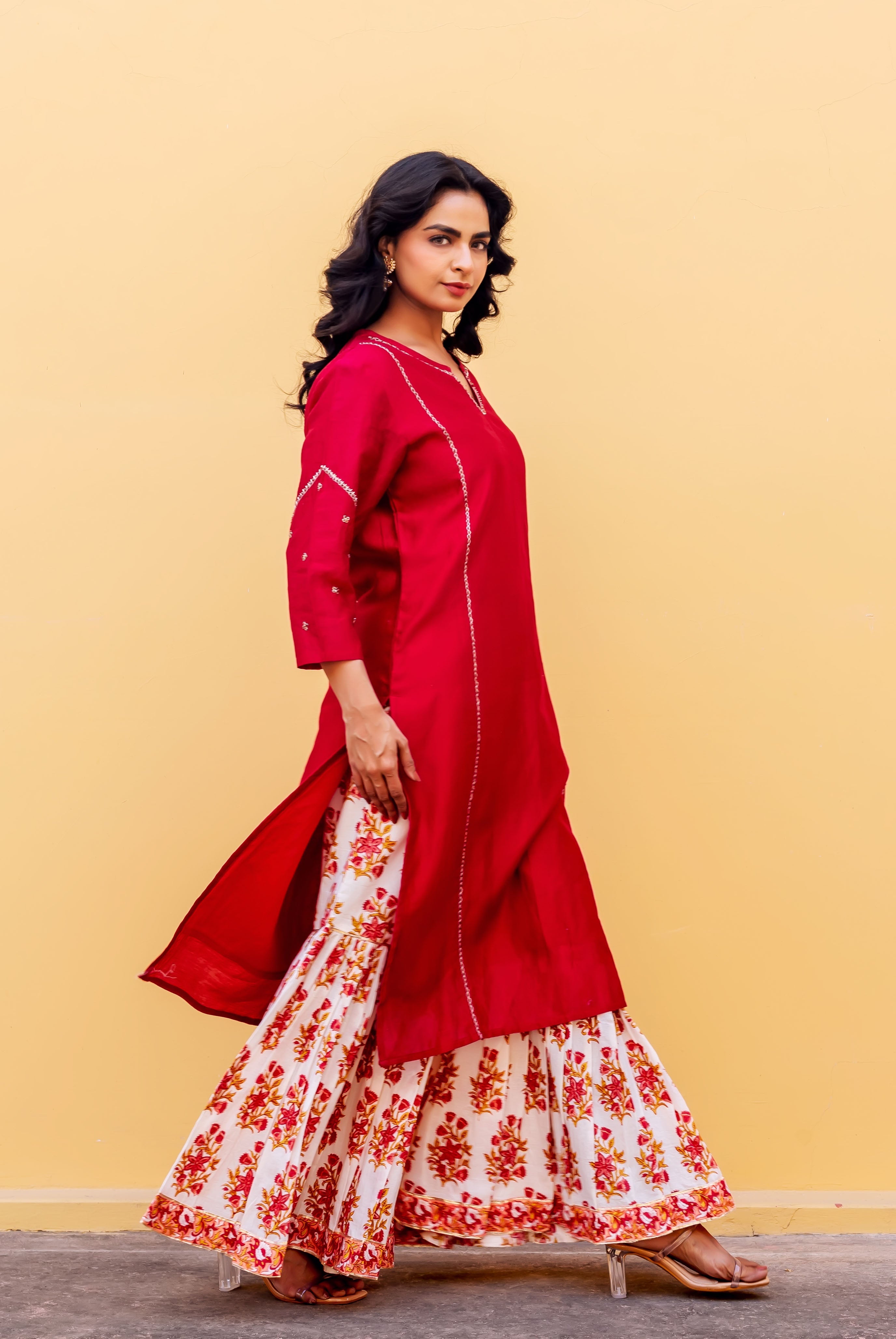 Model in a red embroidered kurta and white floral hand-block printed sharara against a yellow wall.