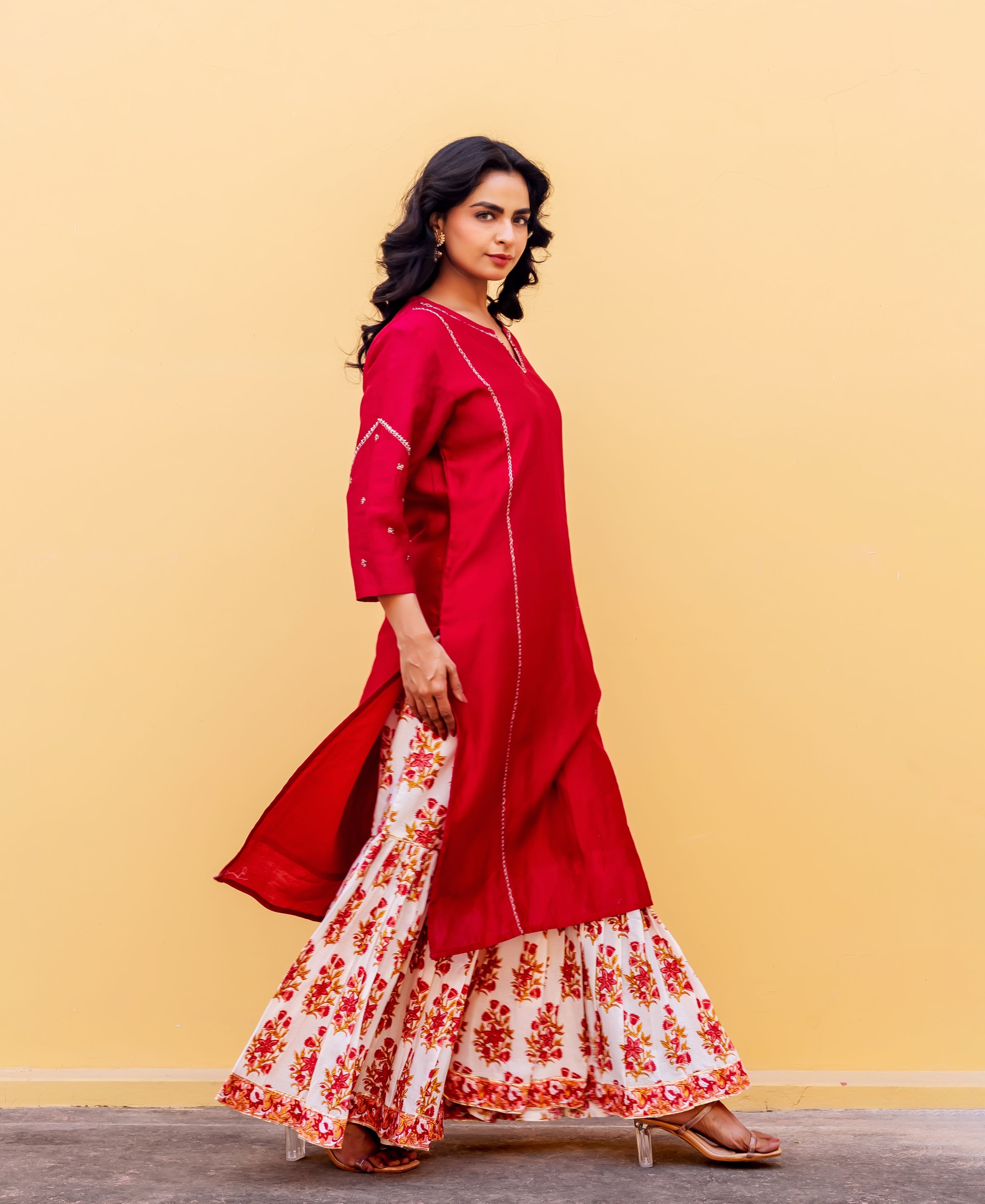 Model in a red embroidered kurta and white floral hand-block printed sharara against a yellow wall.