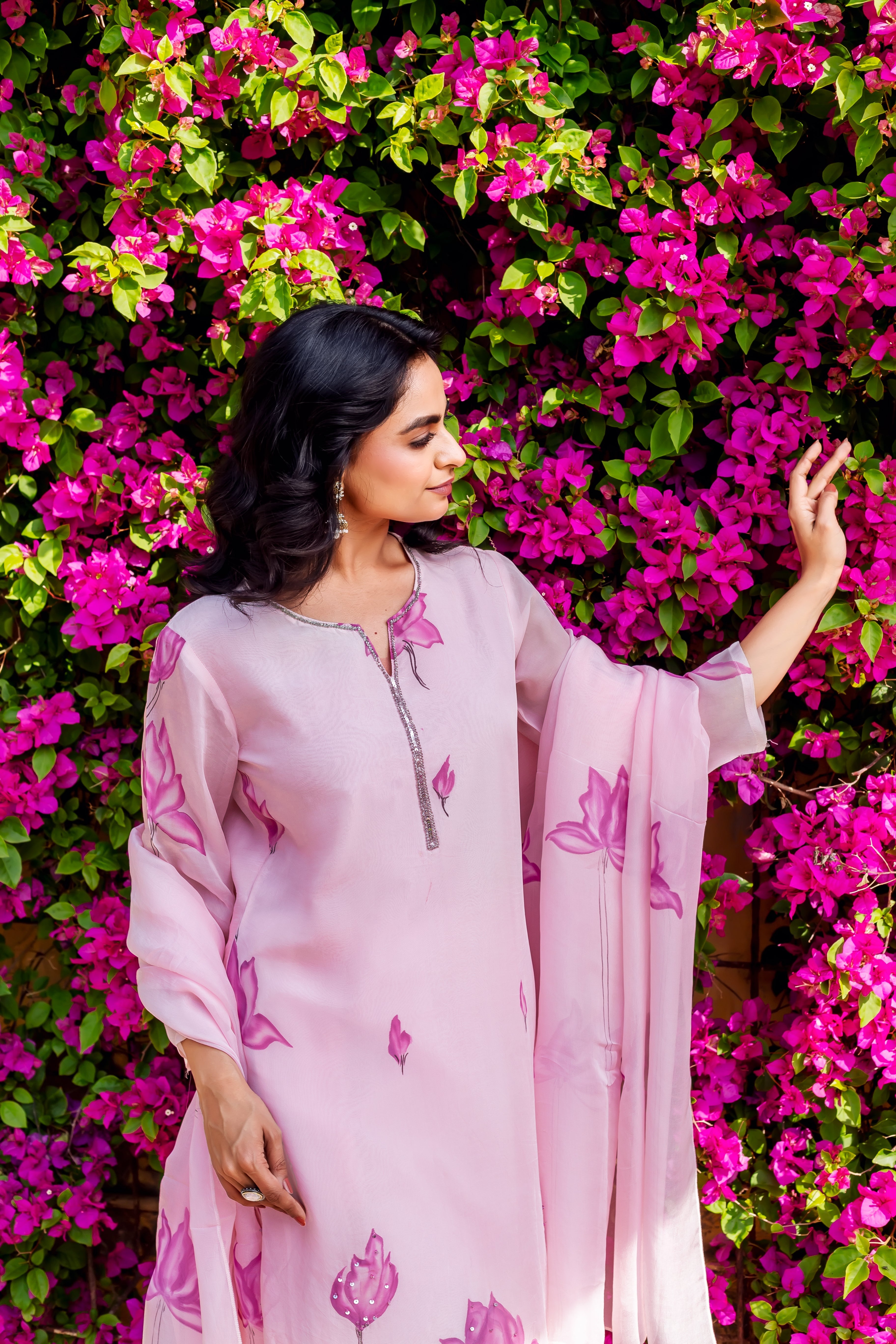 Woman wearing pink organza kurta with darker pink floral print, posing beside magenta flowers.