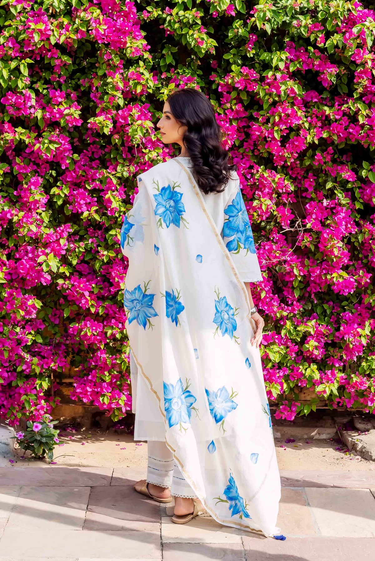 Woman wears white kurta with blue floral brush-painted design, standing before pink flowers.