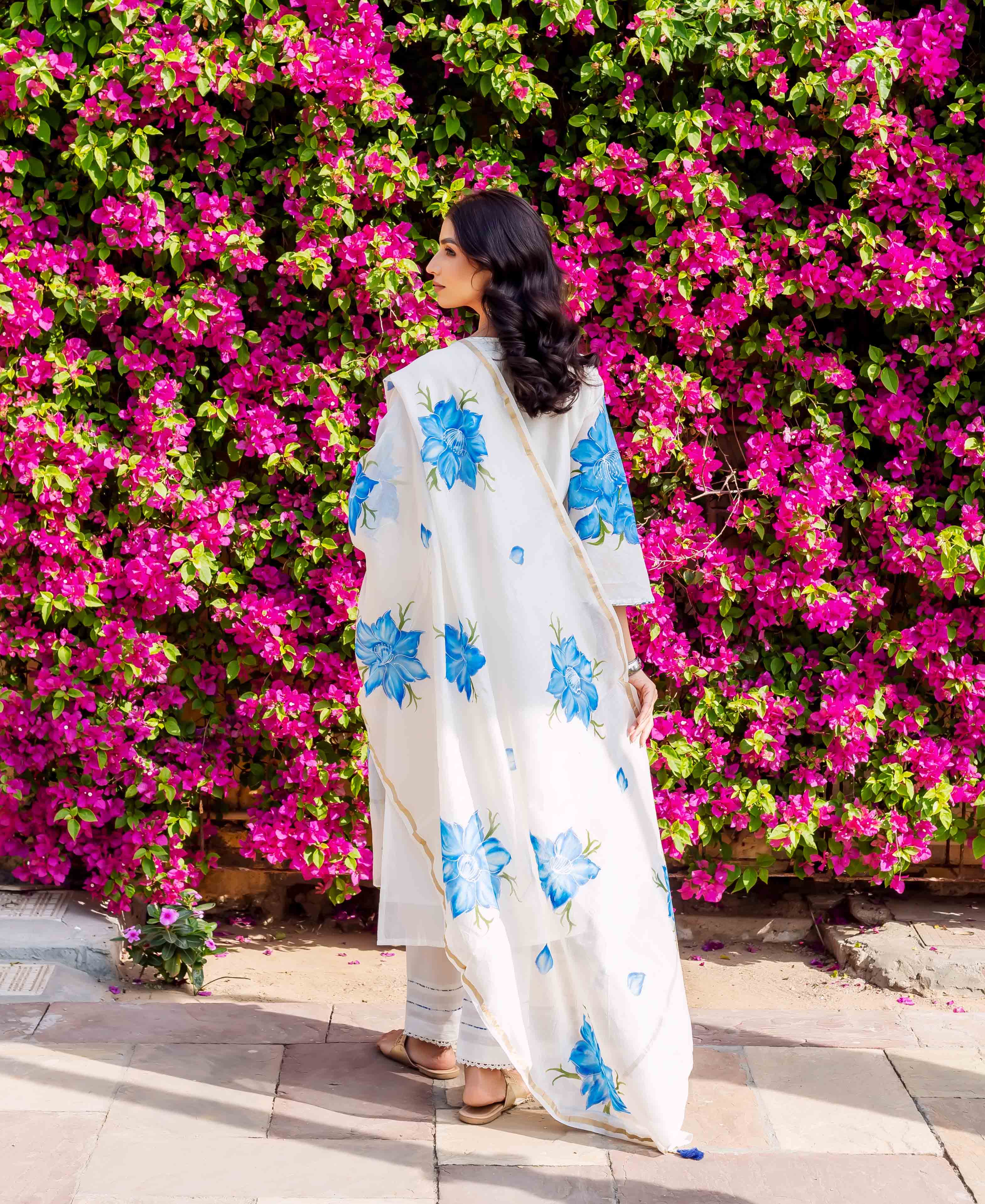 Woman wears white kurta with blue floral brush-painted design, standing before pink flowers.