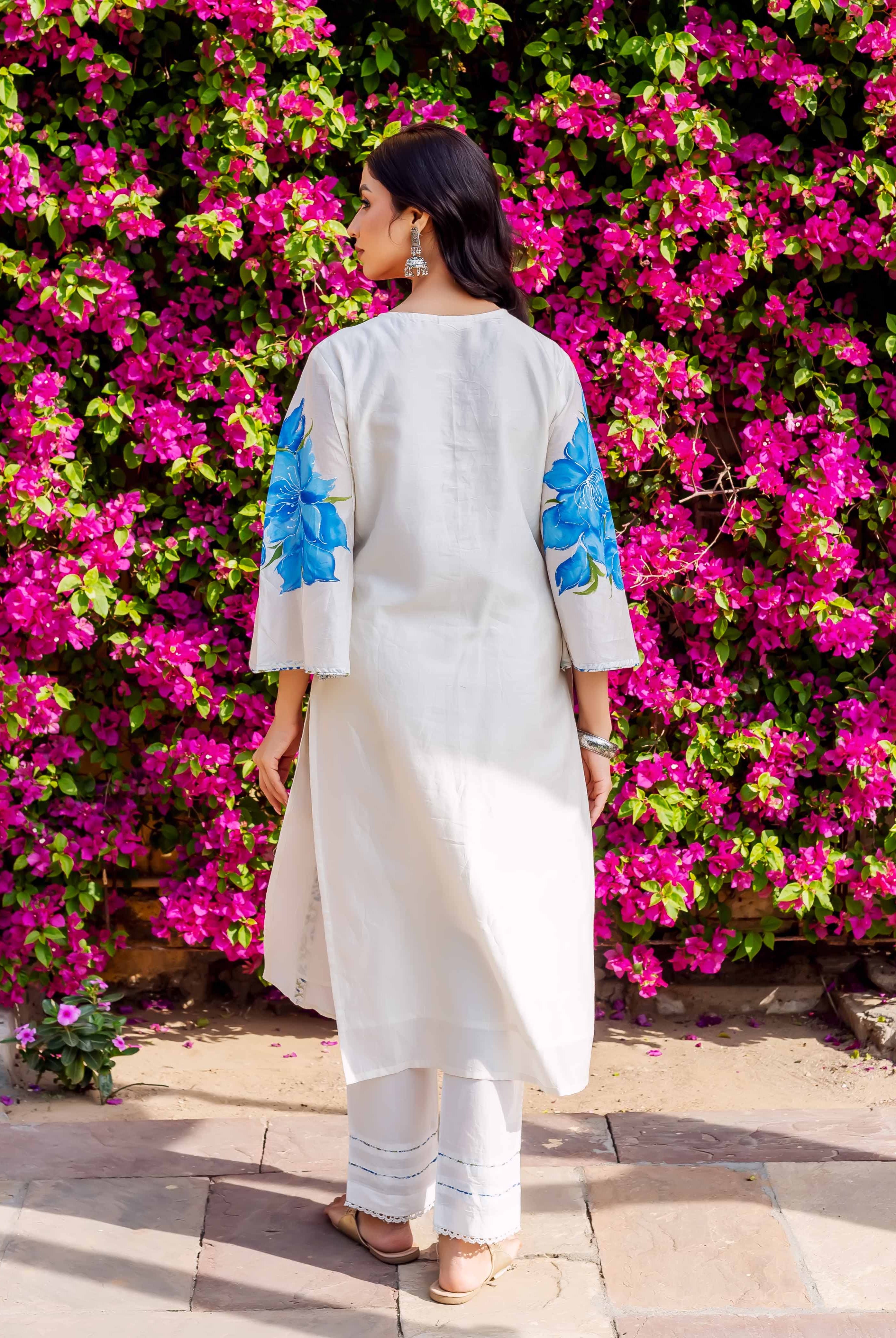 A woman in a white kurta with blue floral sleeves, standing in front of a pink flower wall.