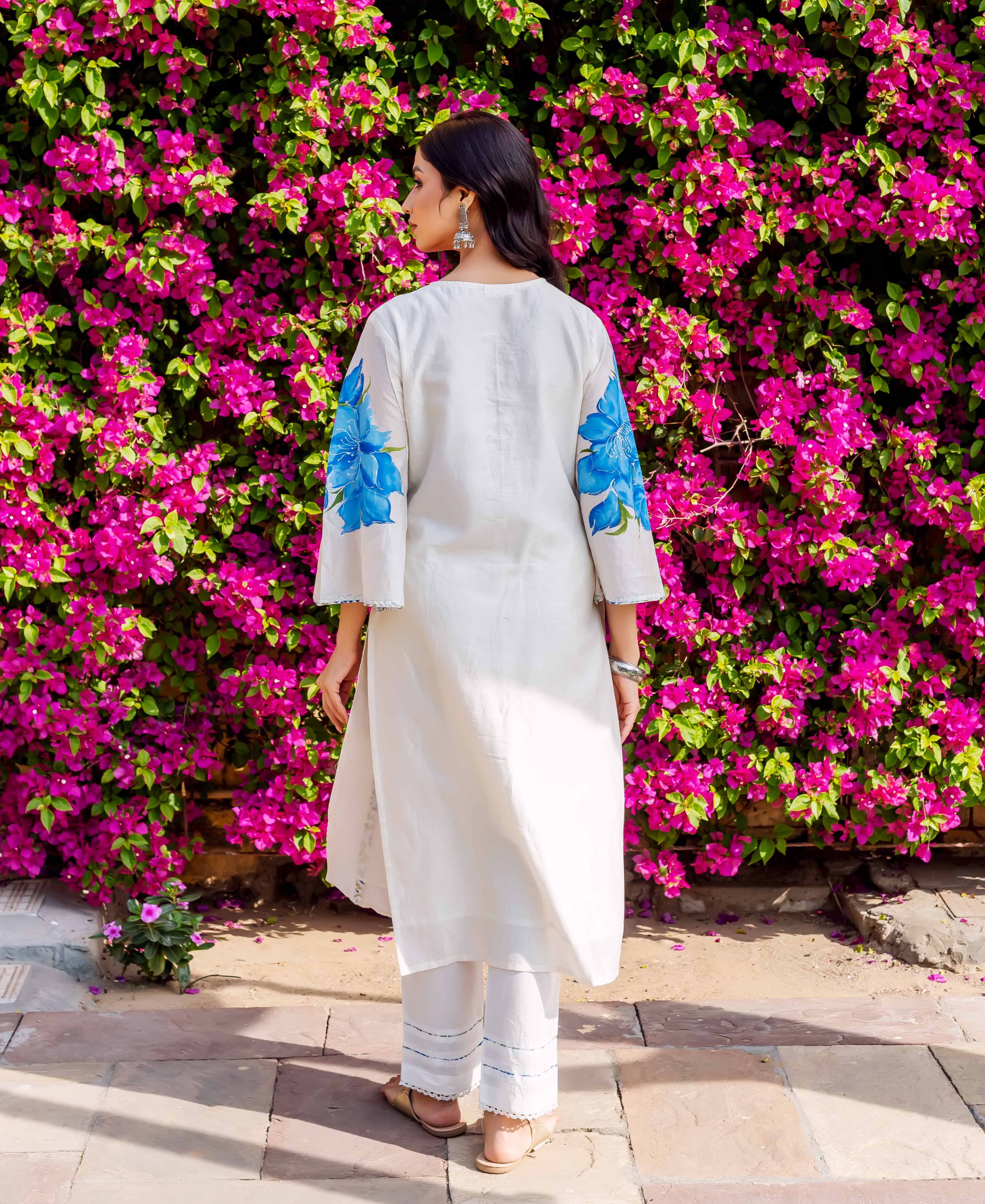 A woman in a white kurta with blue floral sleeves, standing in front of a pink flower wall.
