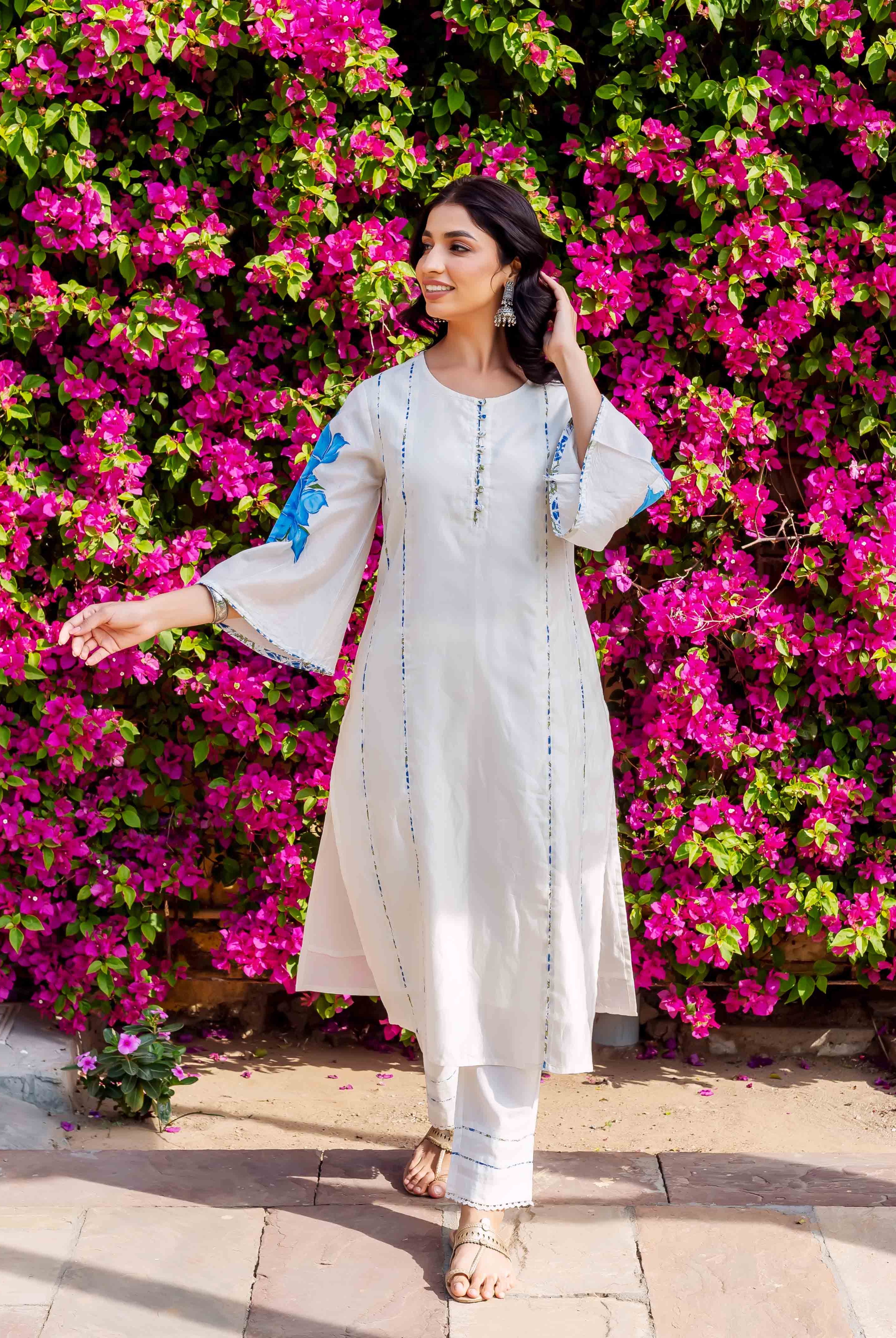 Woman in white kurta and pants with blue brush-painted patterns, posing front against flowers.