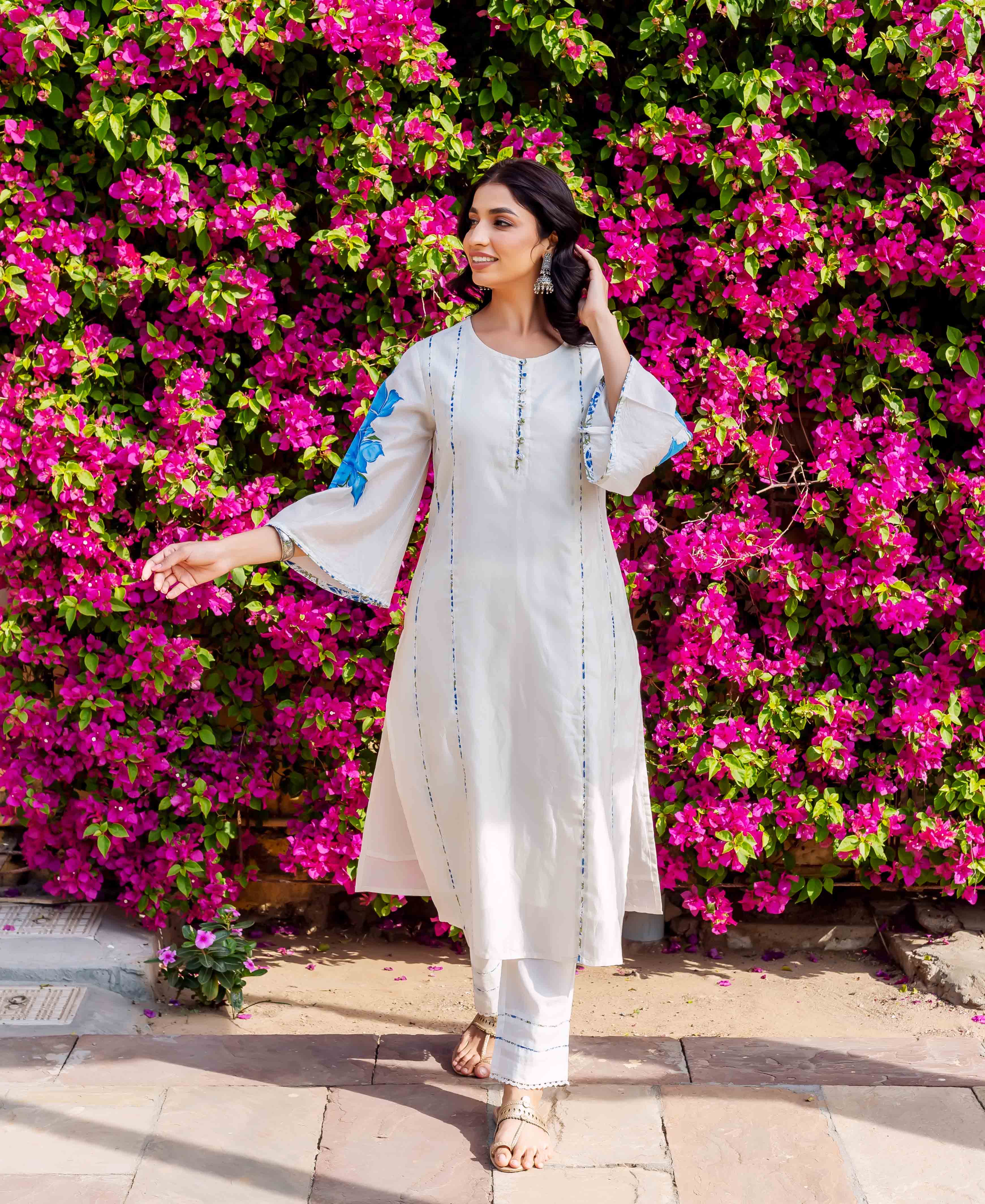 Woman in white kurta and pants with blue brush-painted patterns, posing front against flowers.