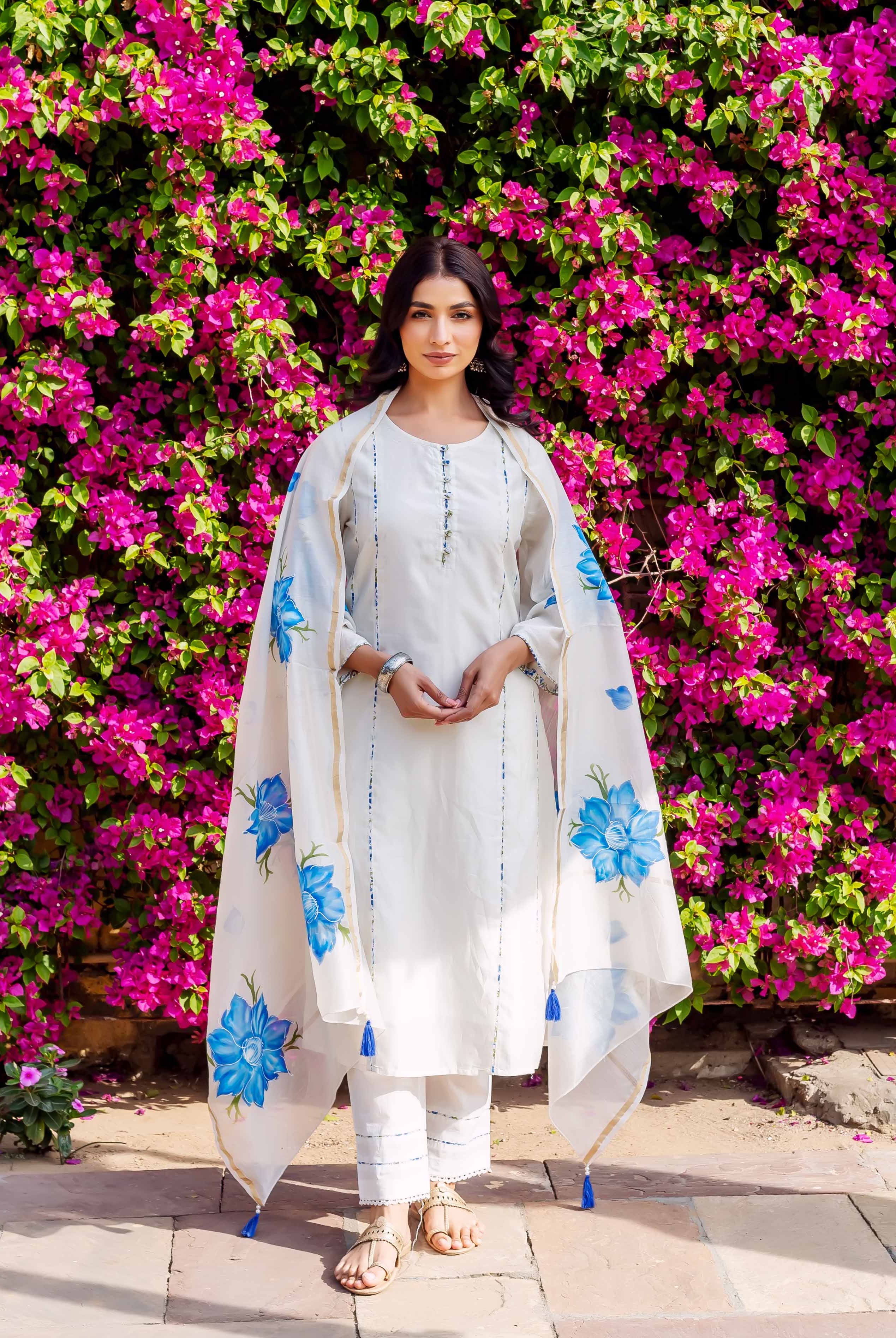 Woman wearing white kurta with blue accents and floral dupatta standing before pink flowers.