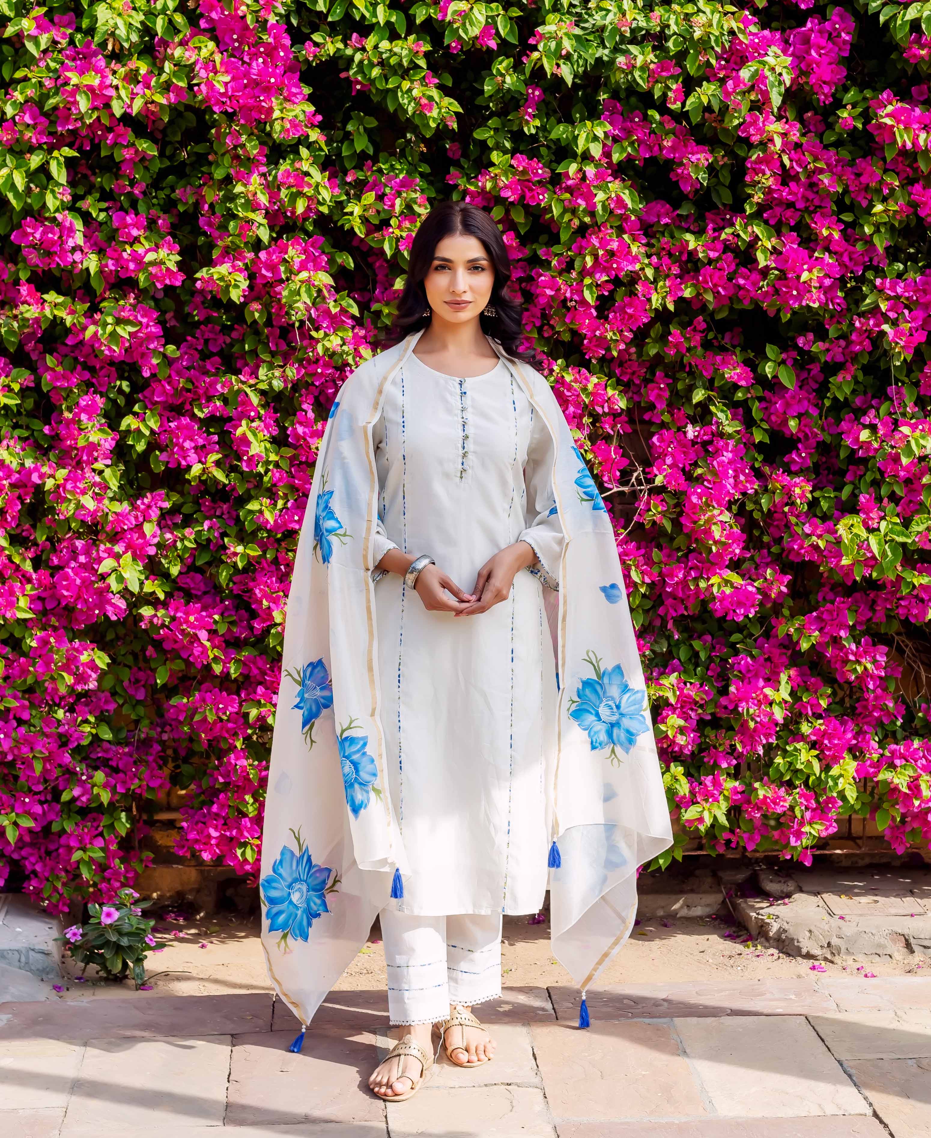 Woman wearing white kurta with blue accents and floral dupatta standing before pink flowers.