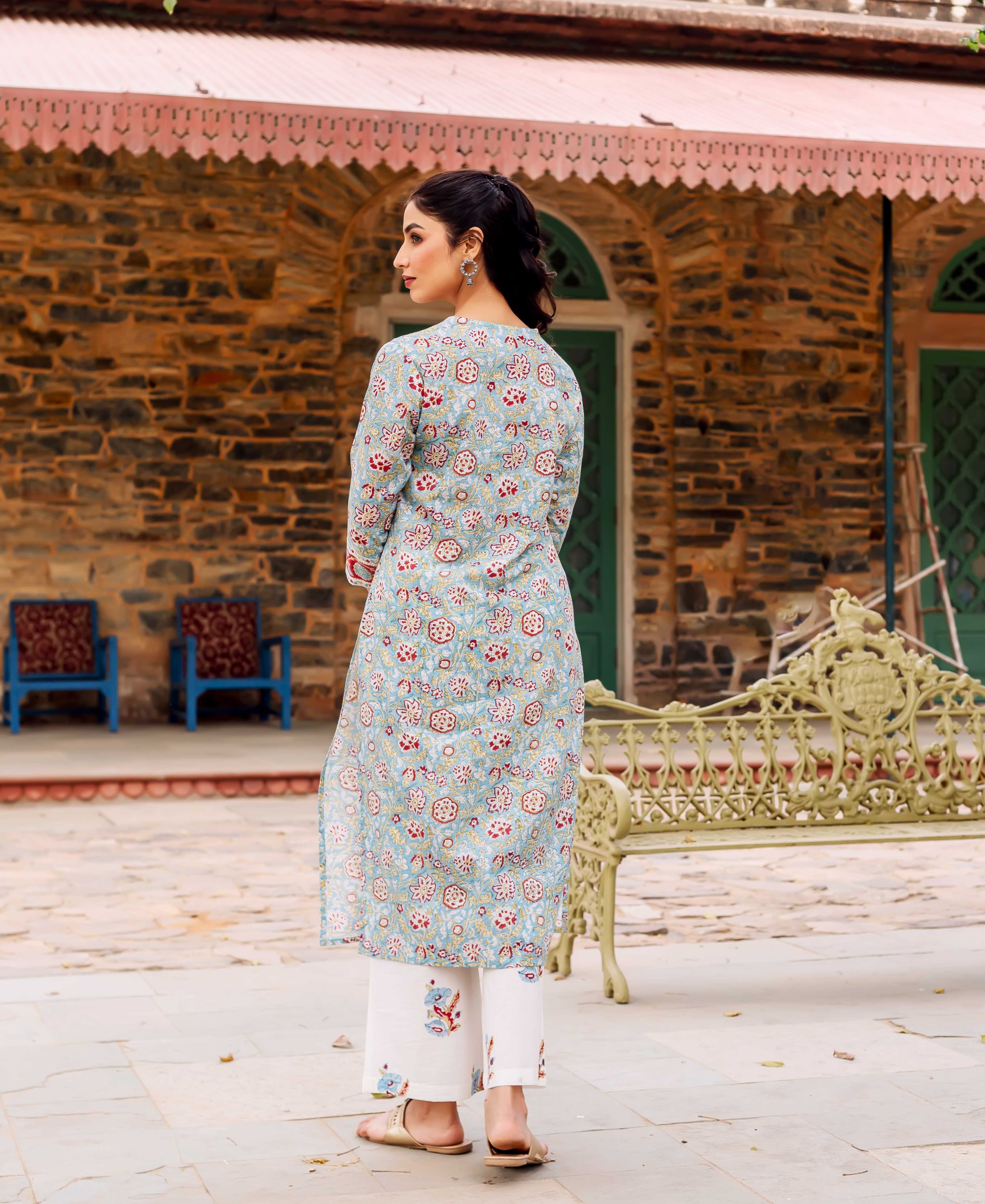 A woman wearing a teal kurta with a floral print, standing in front of a stone building.