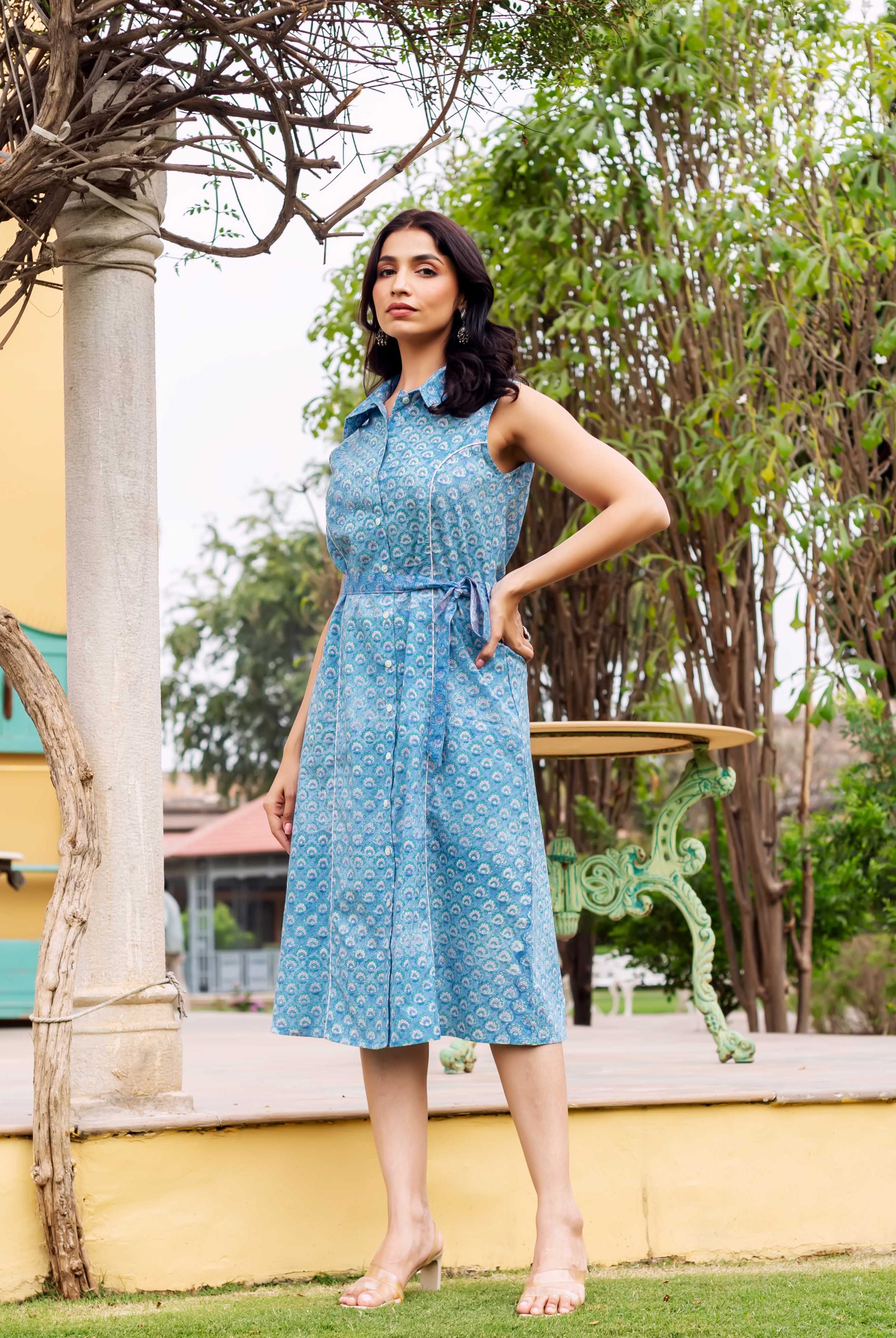 Full-body model wears a blue block-printed sleeveless button-down dress with a matching belt.