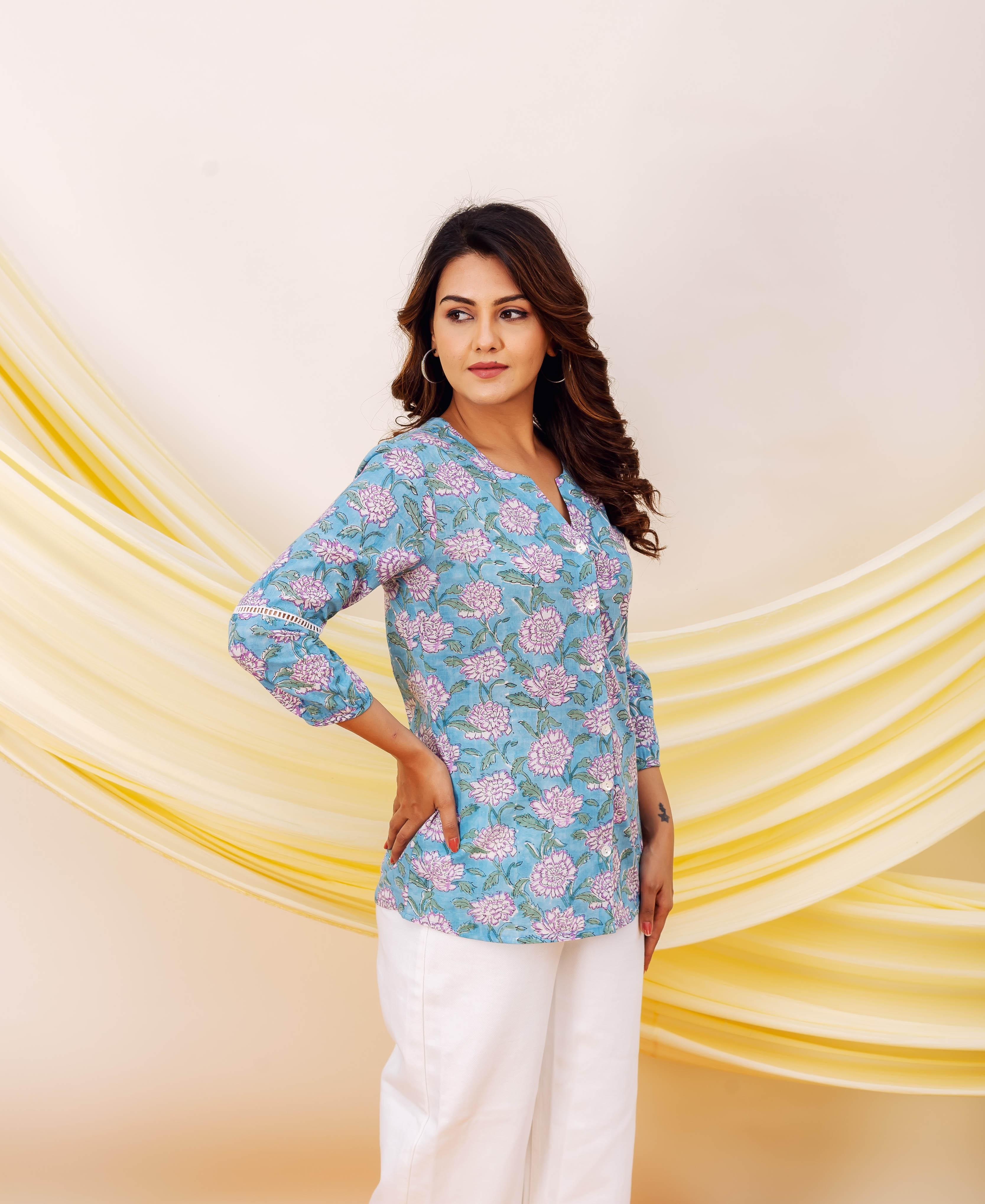 Model posing in a sky blue floral printed top with white lace trim and white pants.