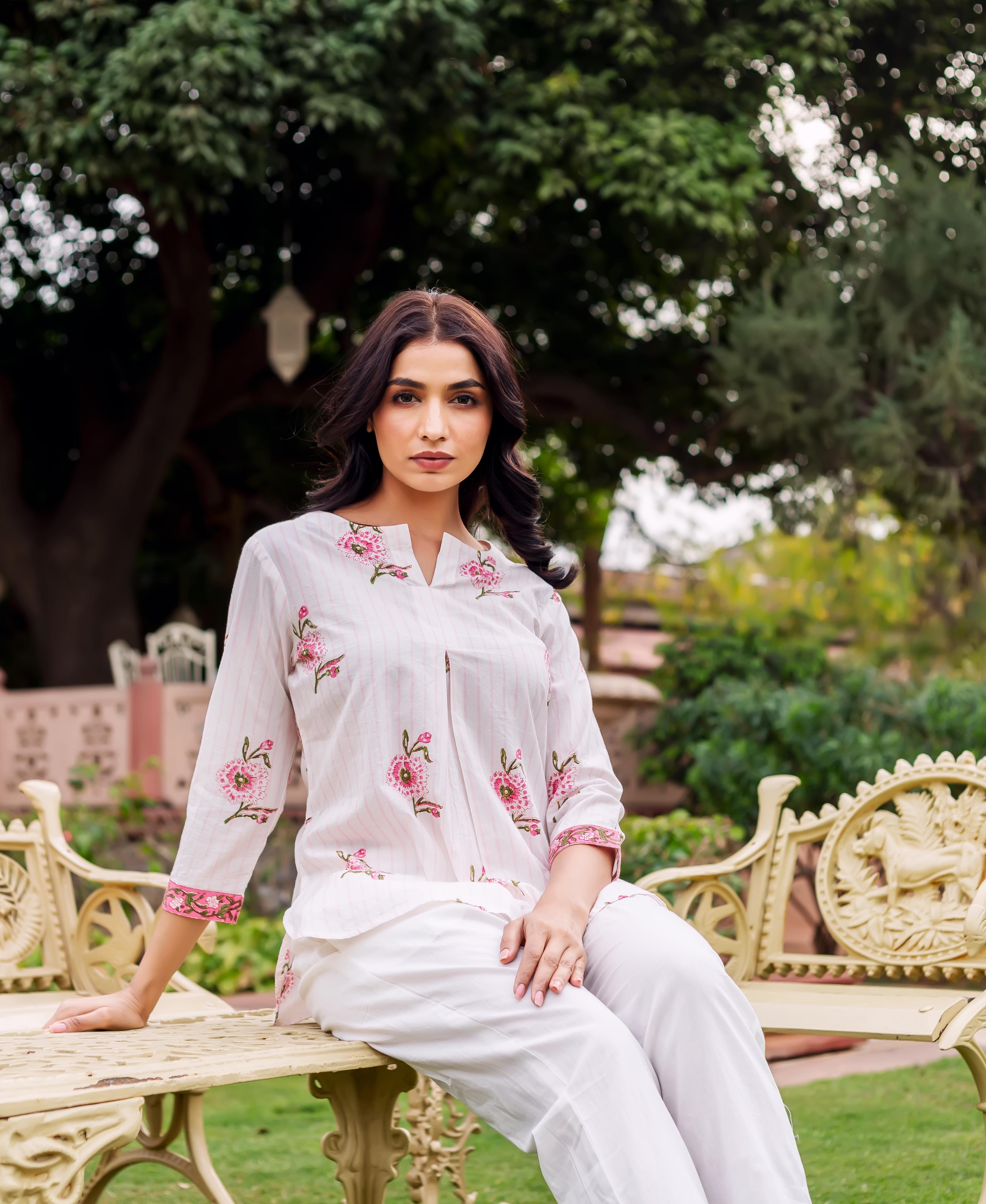 Woman in white tunic with pink floral embroidery and white pants sitting on beige bench.