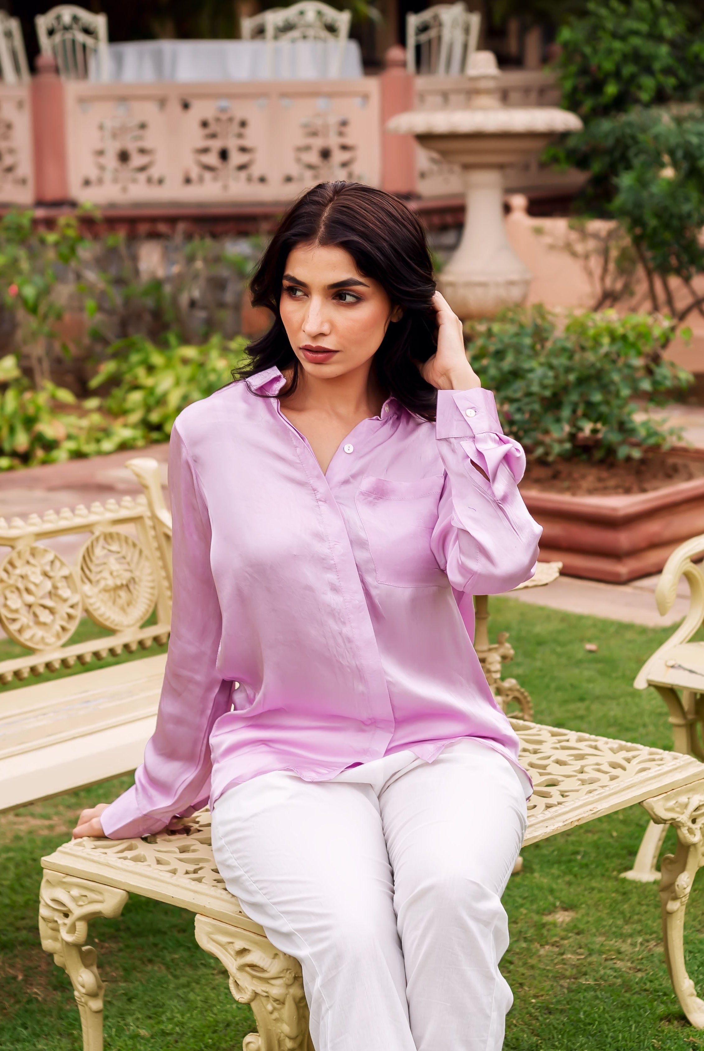 Woman wearing a pastel pink button-down shirt and white pants sitting on a bench.