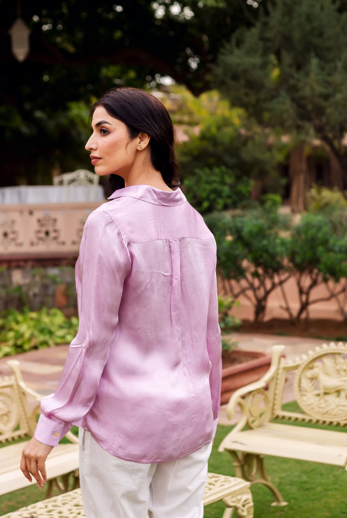 A woman wearing a pink button-down shirt with a subtle sheen, standing in a garden setting.