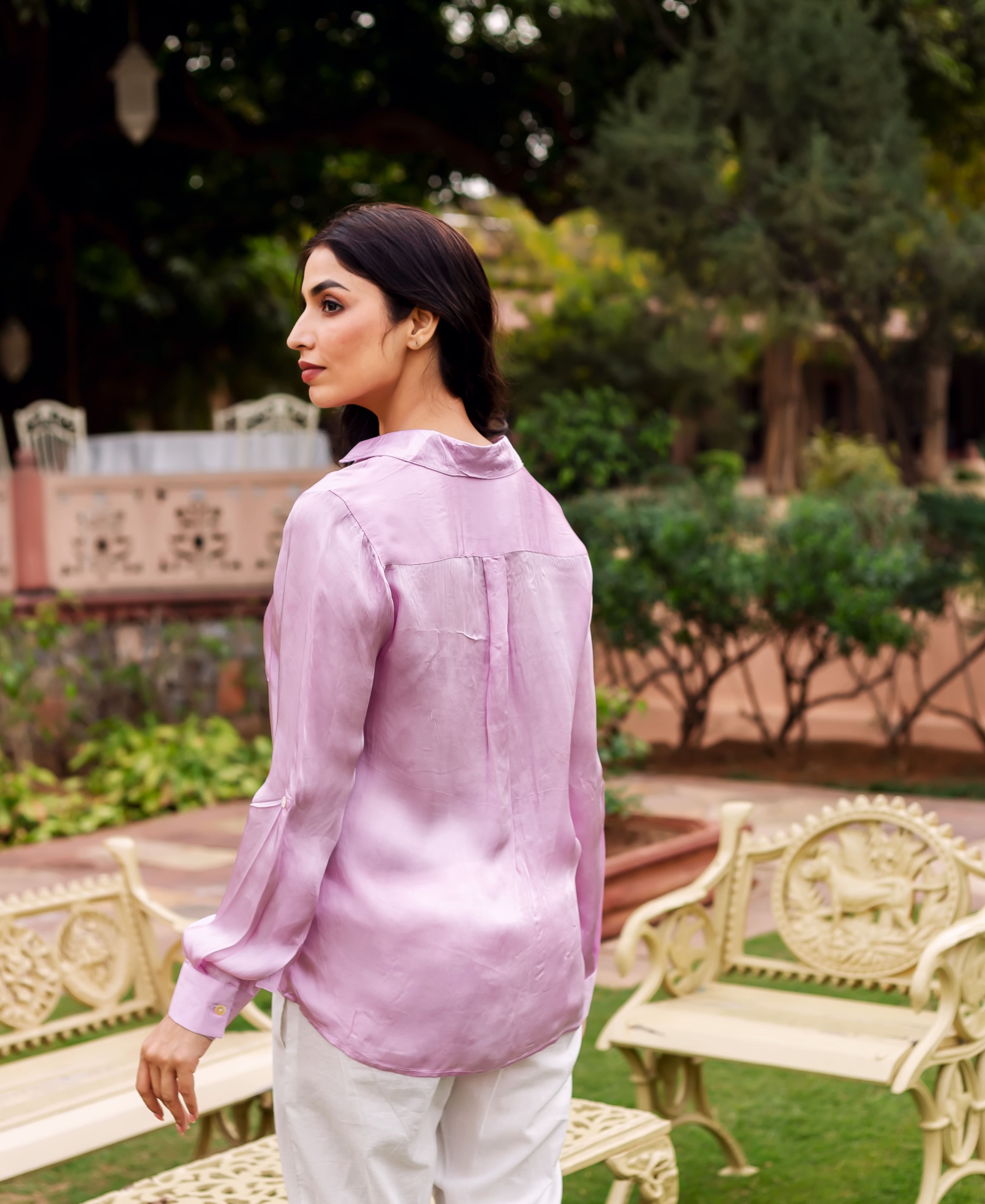 A woman wearing a pink button-down shirt with a subtle sheen, standing in a garden setting.