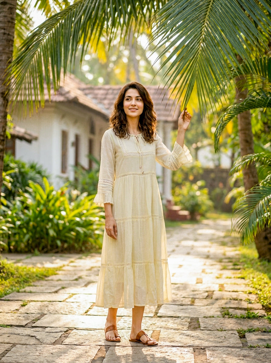 A woman in a long, ivory, tiered cotton dress with lace inserts and brown sandals.