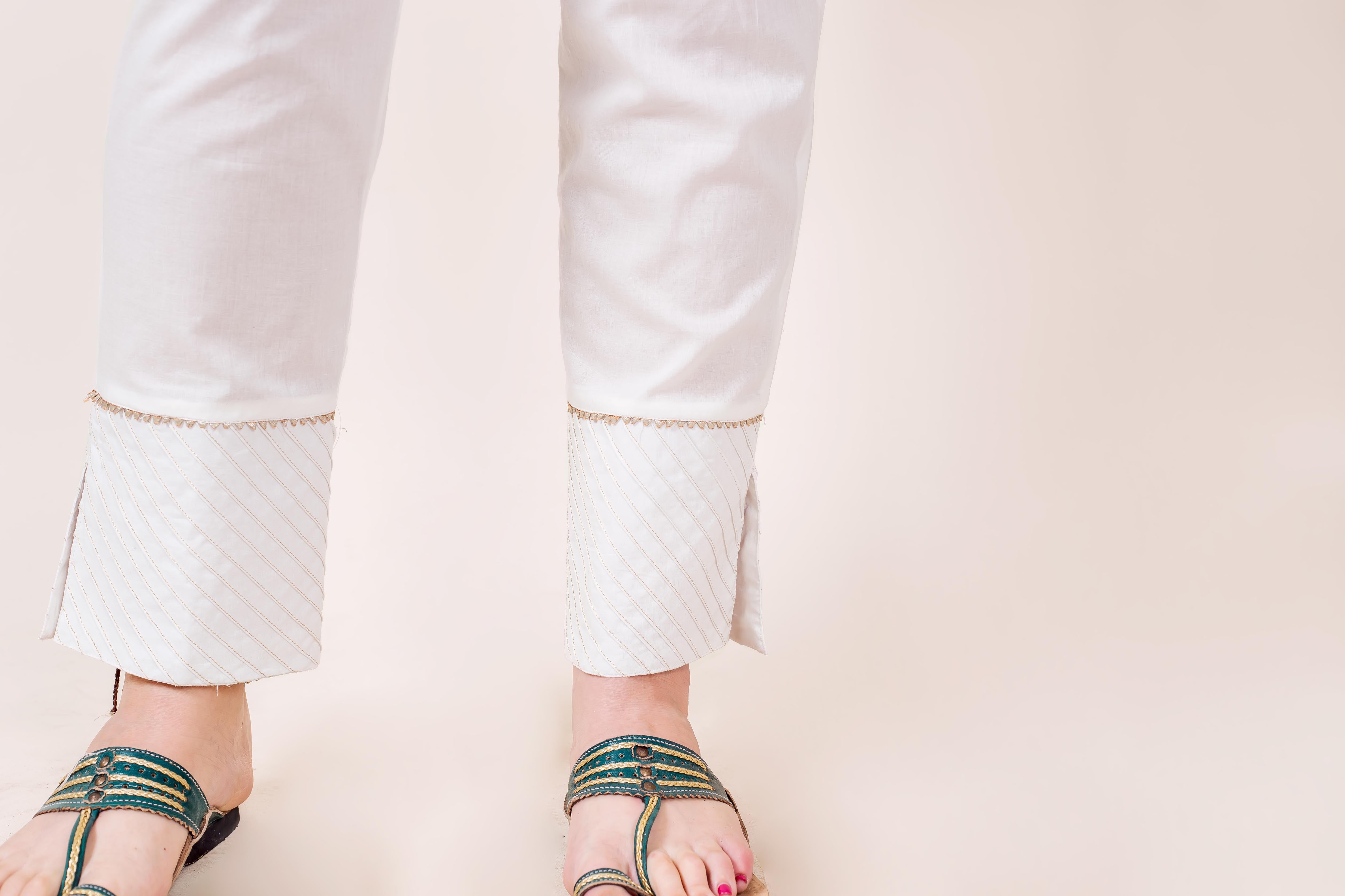 White straight pants on a model, featuring golden trim and striped cuffs with slits.