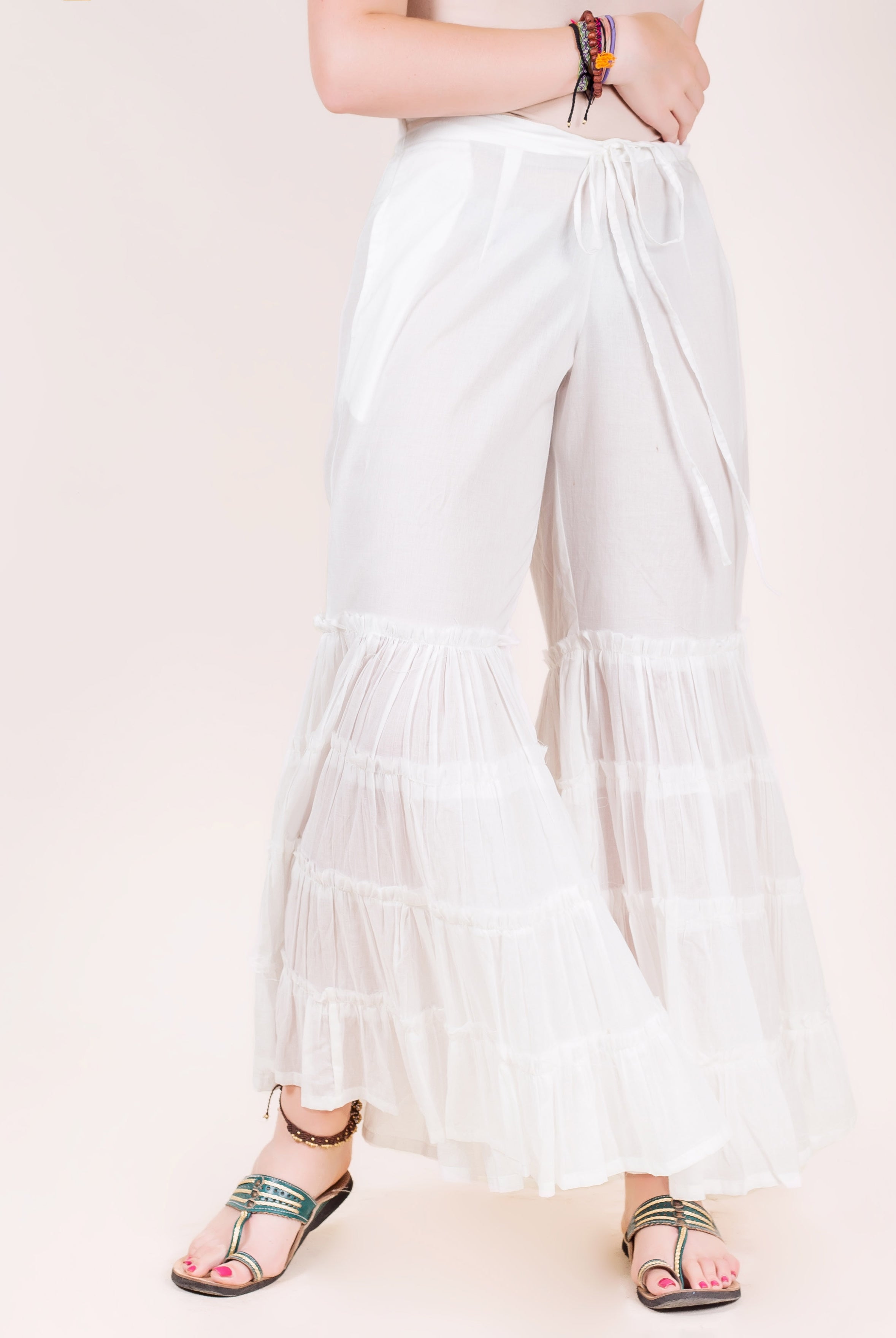 White tiered sharara pants with ruffled hem, on model with sandals and colorful bracelets.