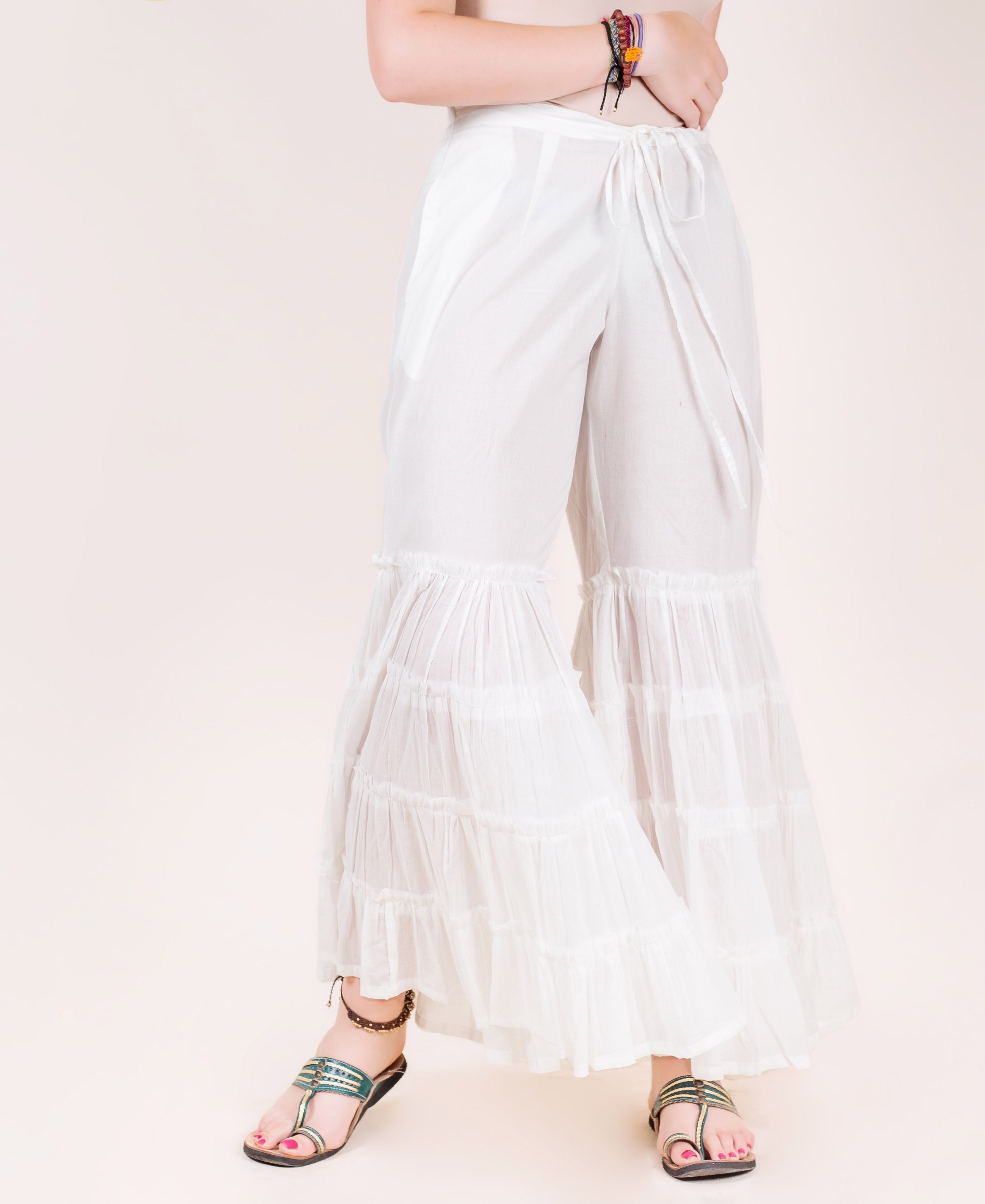 White tiered sharara pants with ruffled hem, on model with sandals and colorful bracelets.