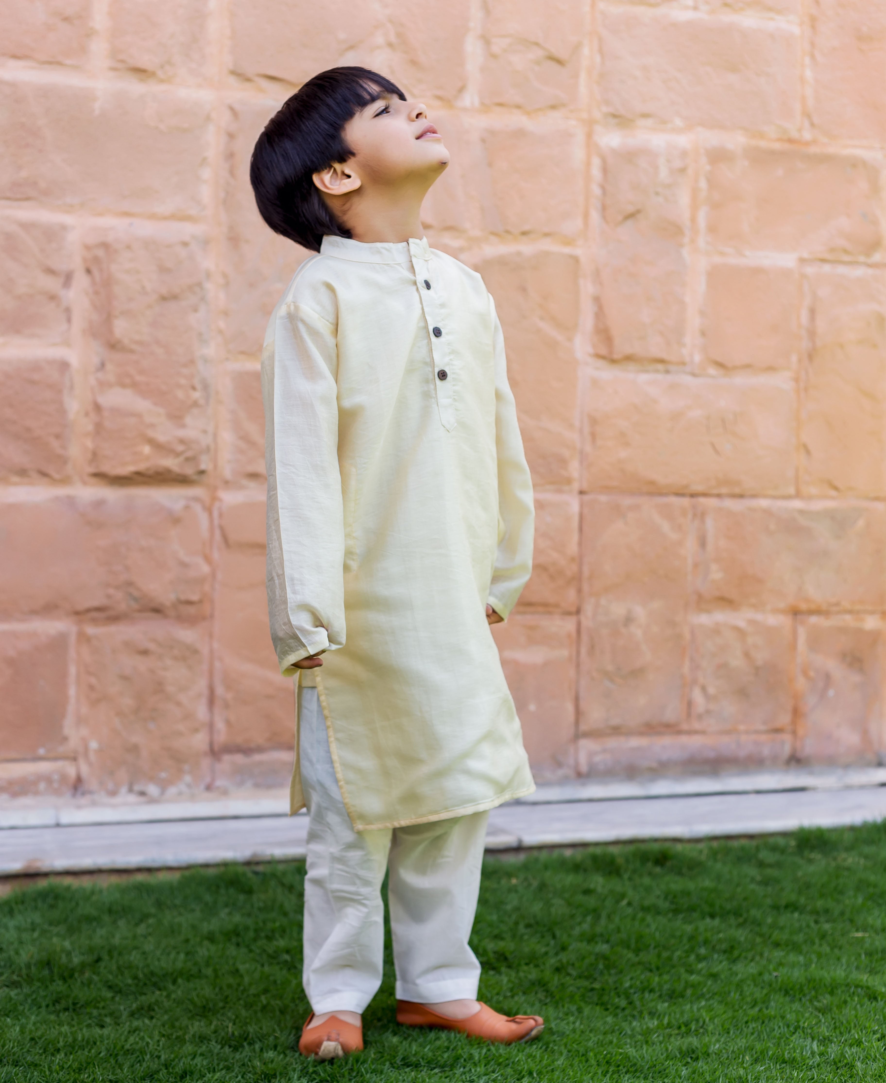 A young boy in a cream-colored kurta pajama set, standing on grass.