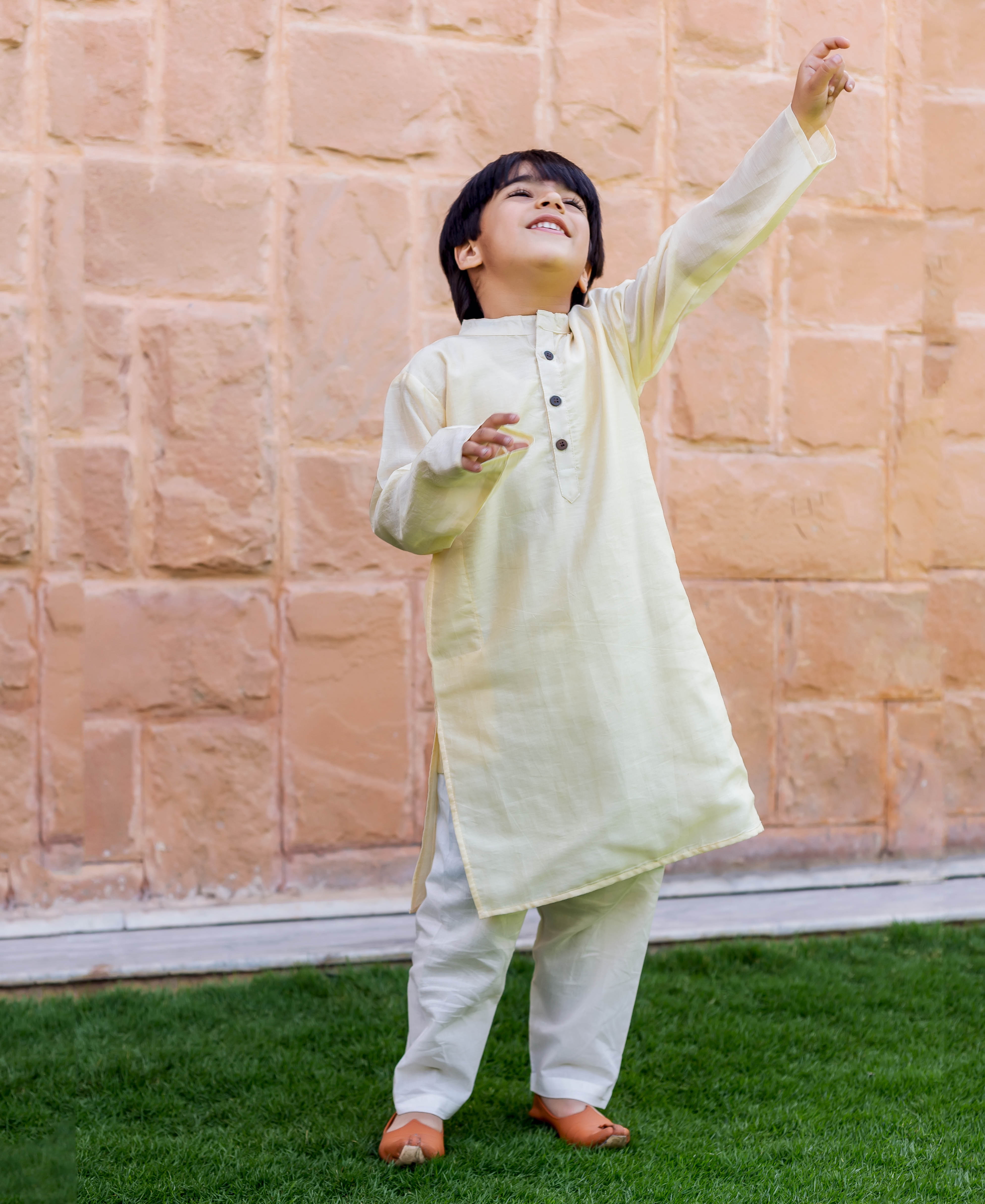 Young boy wearing a crème kurta and white pajama set, looking up and reaching.