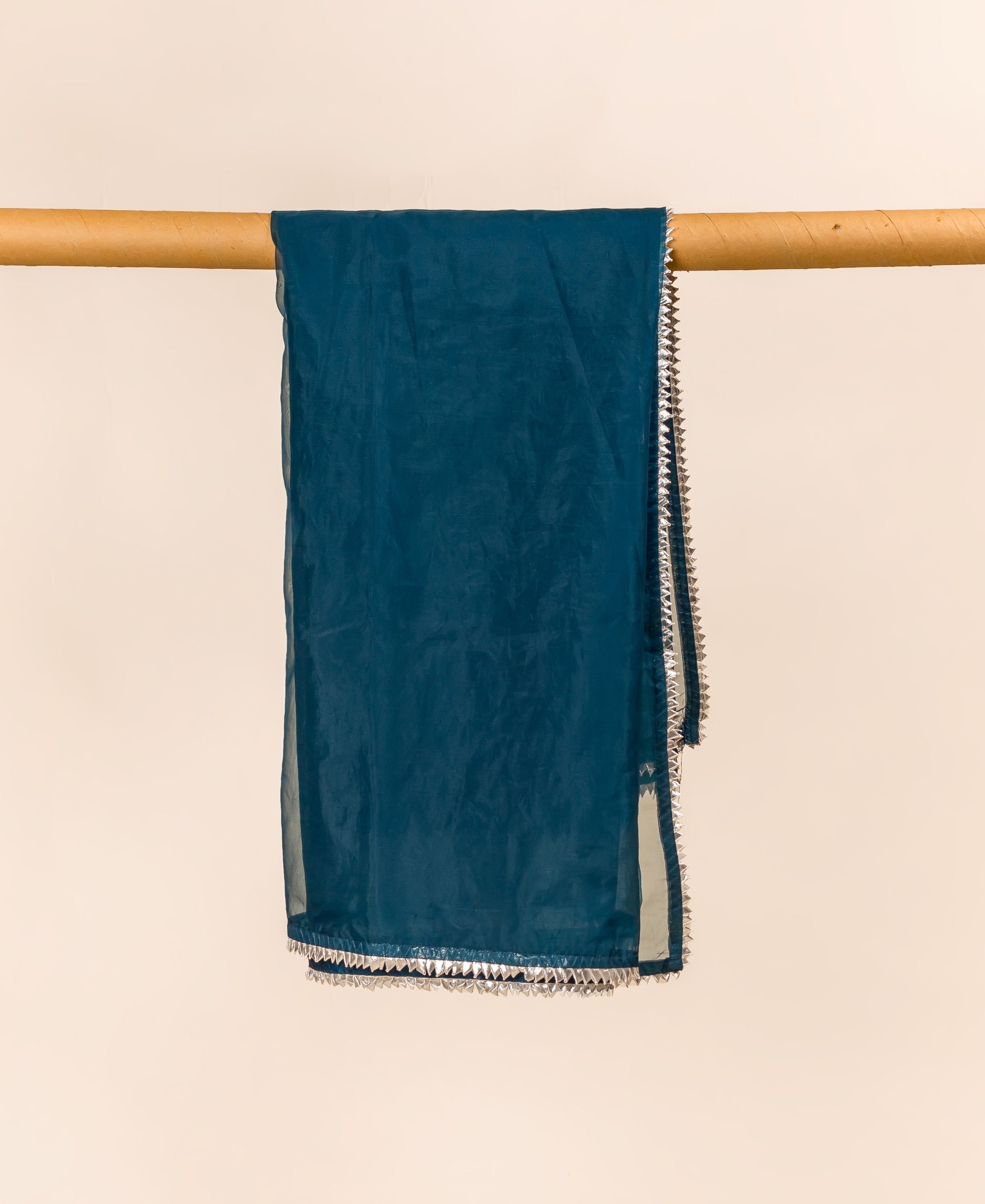 Teal blue organza dupatta with silver fringe, hanging from a wooden rod.