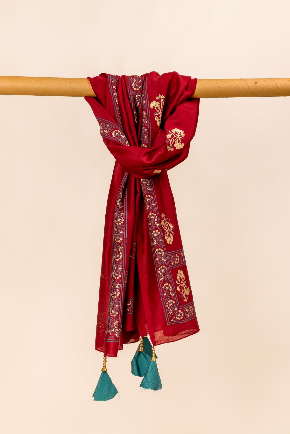 Maroon dupatta draped over a wooden rod, featuring gold block prints and teal tassels.