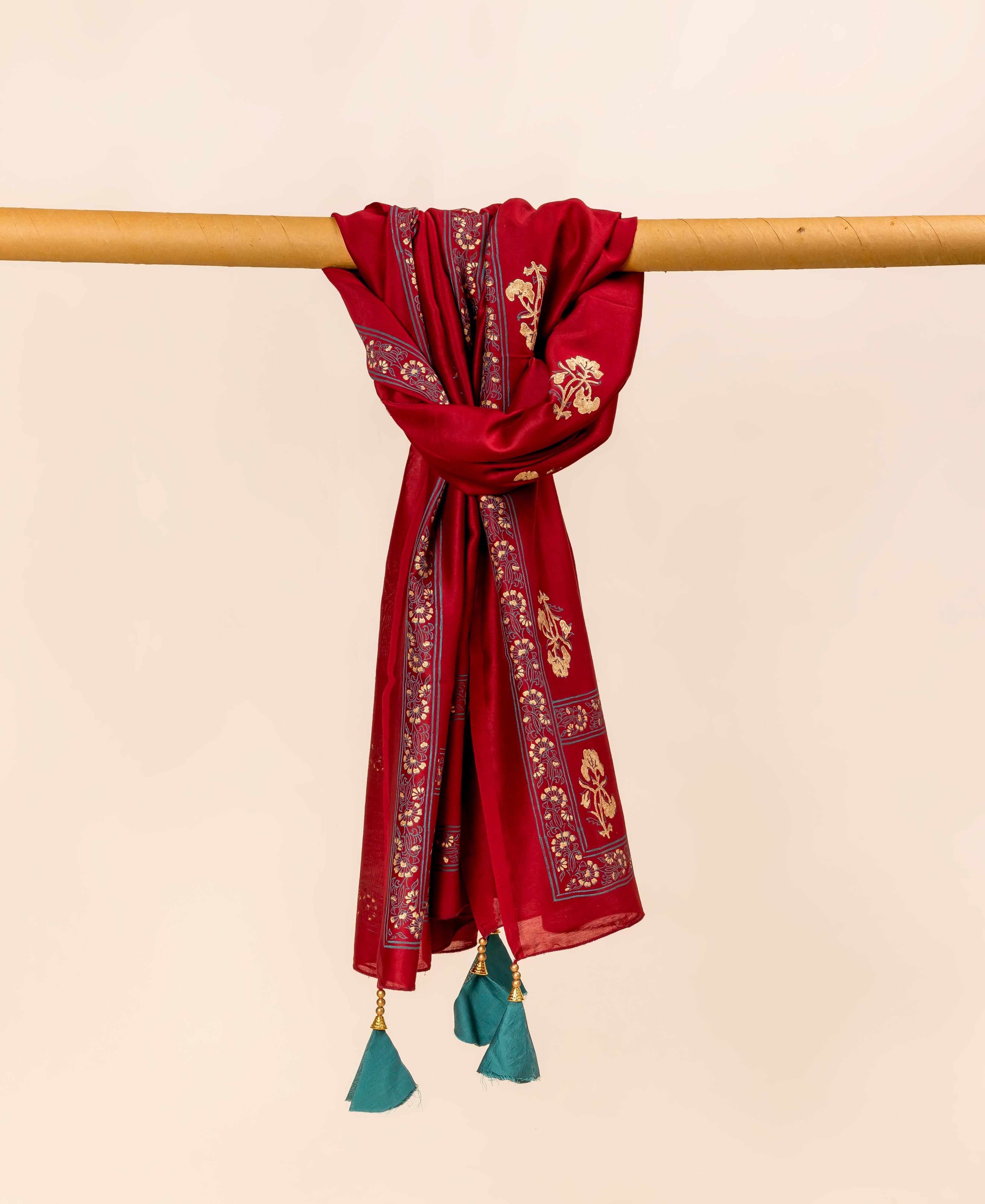 Maroon dupatta draped over a wooden rod, featuring gold block prints and teal tassels.
