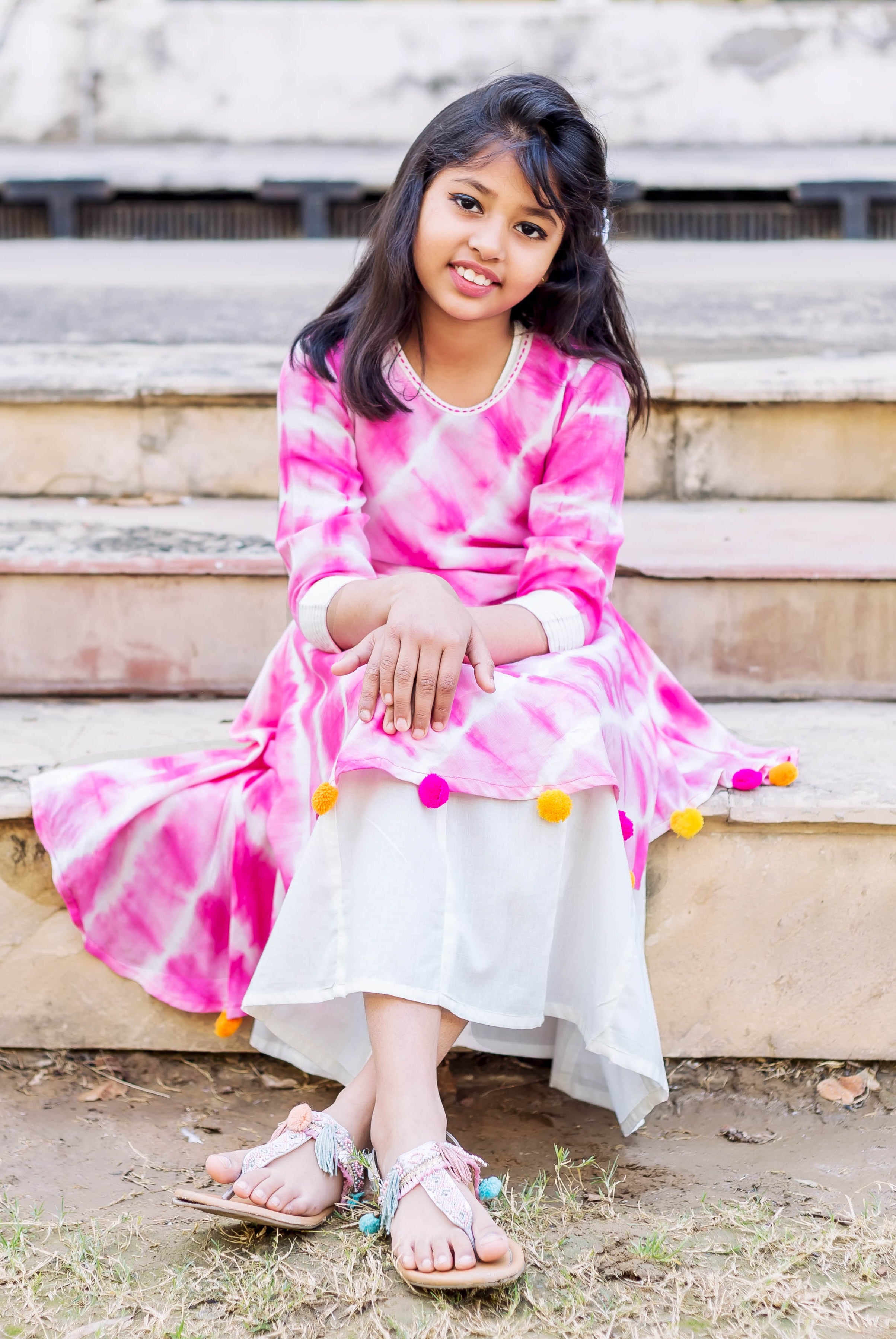 Front view of young model in pink tie-dye asymmetrical dress with white underlayer, seated.